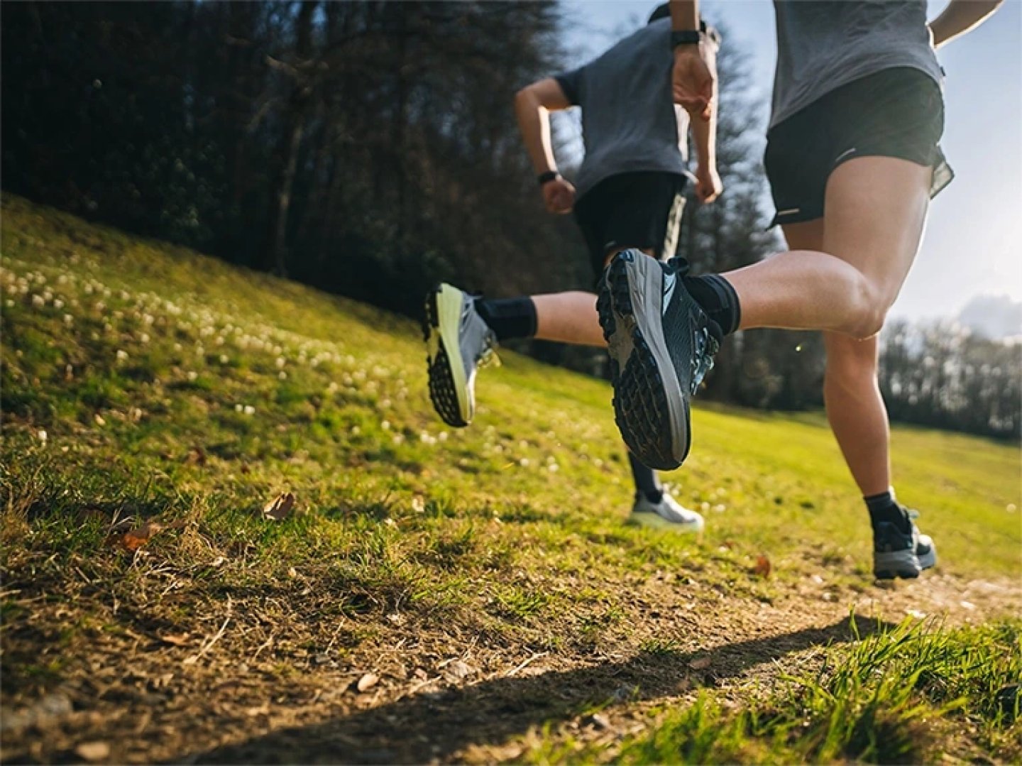 Low-angle action shot of two runners wearing Altra Experience shoes on a grassy dirt trail in morning sunlight.