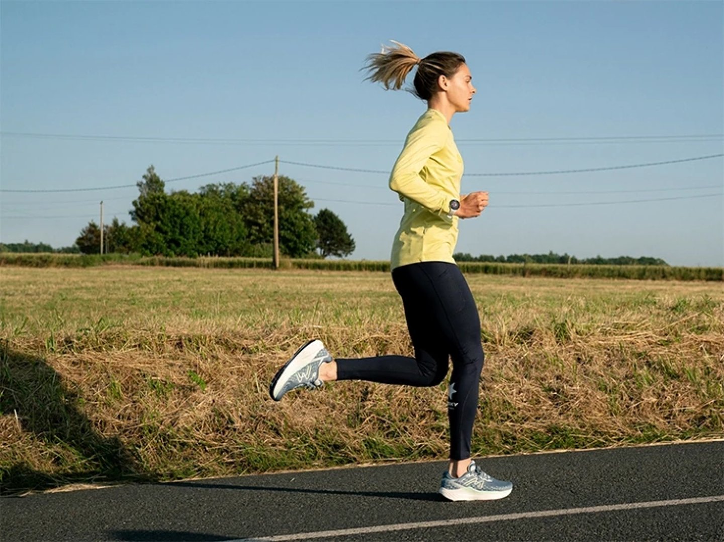 Woman running on a paved rural road wearing a yellow long-sleeve top, black tights, and light blue running shoes