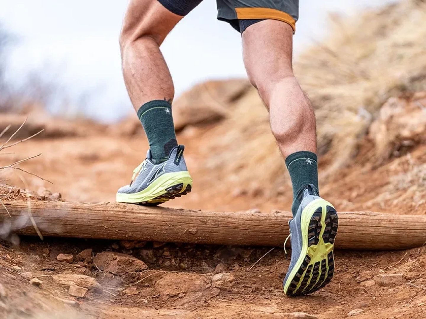 Side profile of Altra trail running shoes with yellow-green outsole worn by a runner stepping over a log on a red dirt trail.