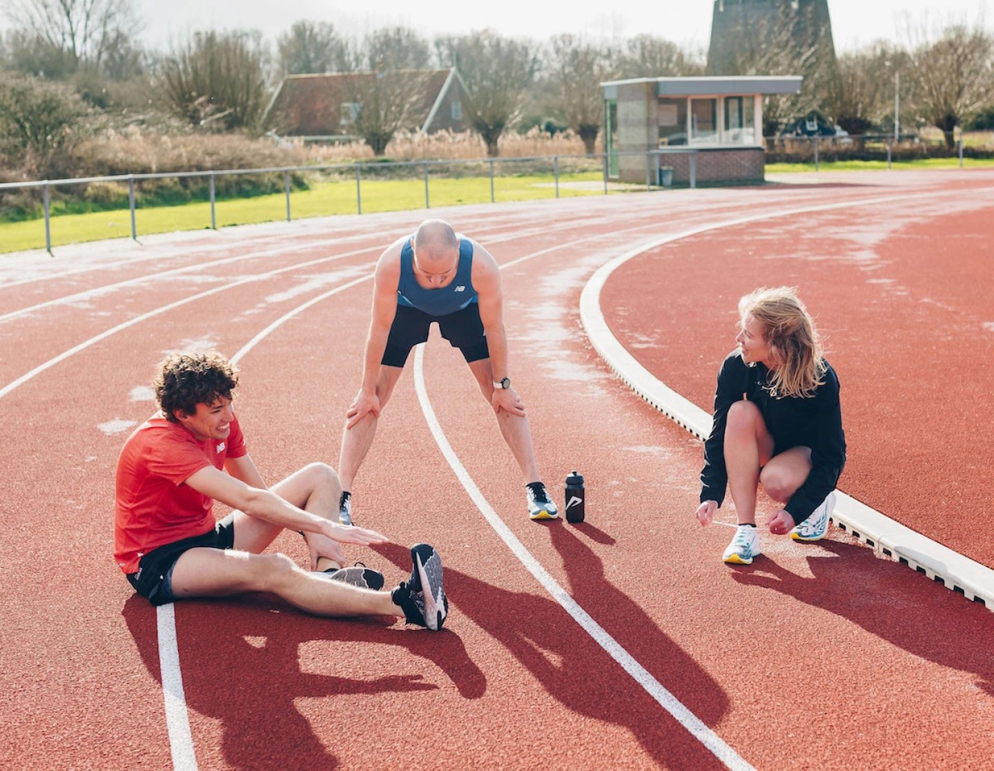 Three runners resting and stretching on a red athletics track, wearing New Balance apparel and running shoes, with a windmill in background.