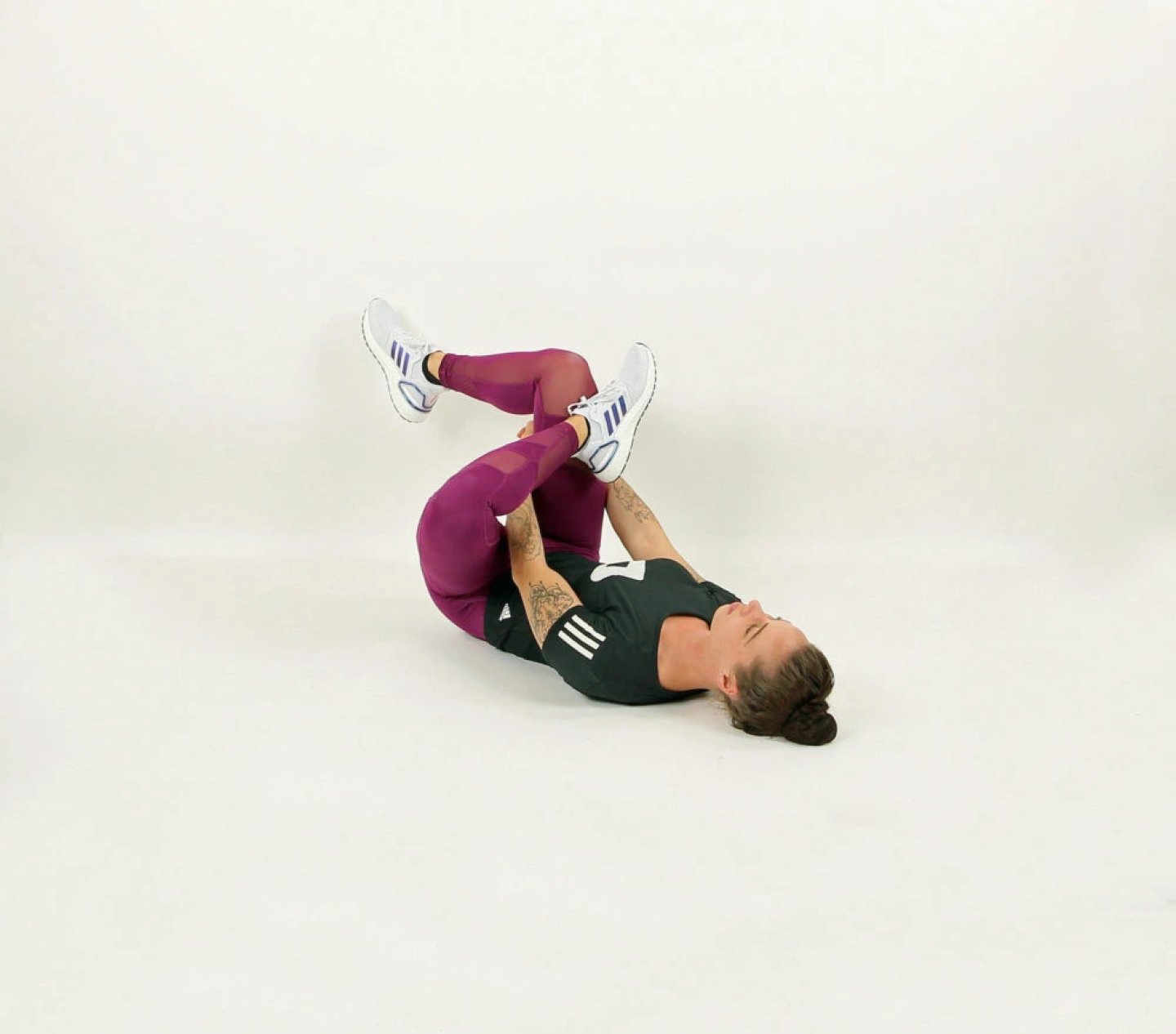 Woman lying on her back pulling both knees to her chest in a supine knee-hug stretch, wearing burgundy leggings and a dark top.