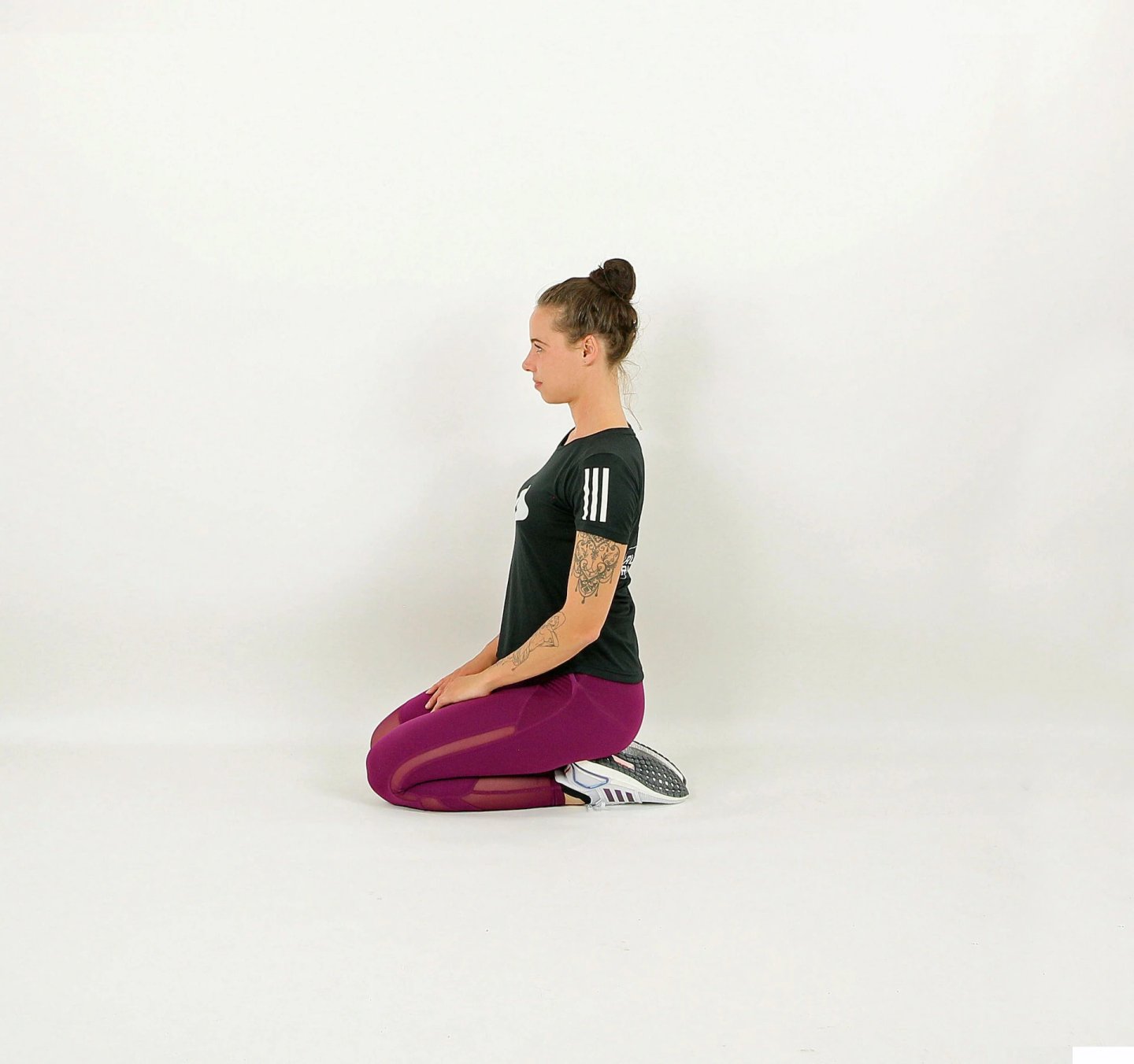 Woman in purple leggings and black Adidas t-shirt kneeling upright on floor performing a quad stretch exercise