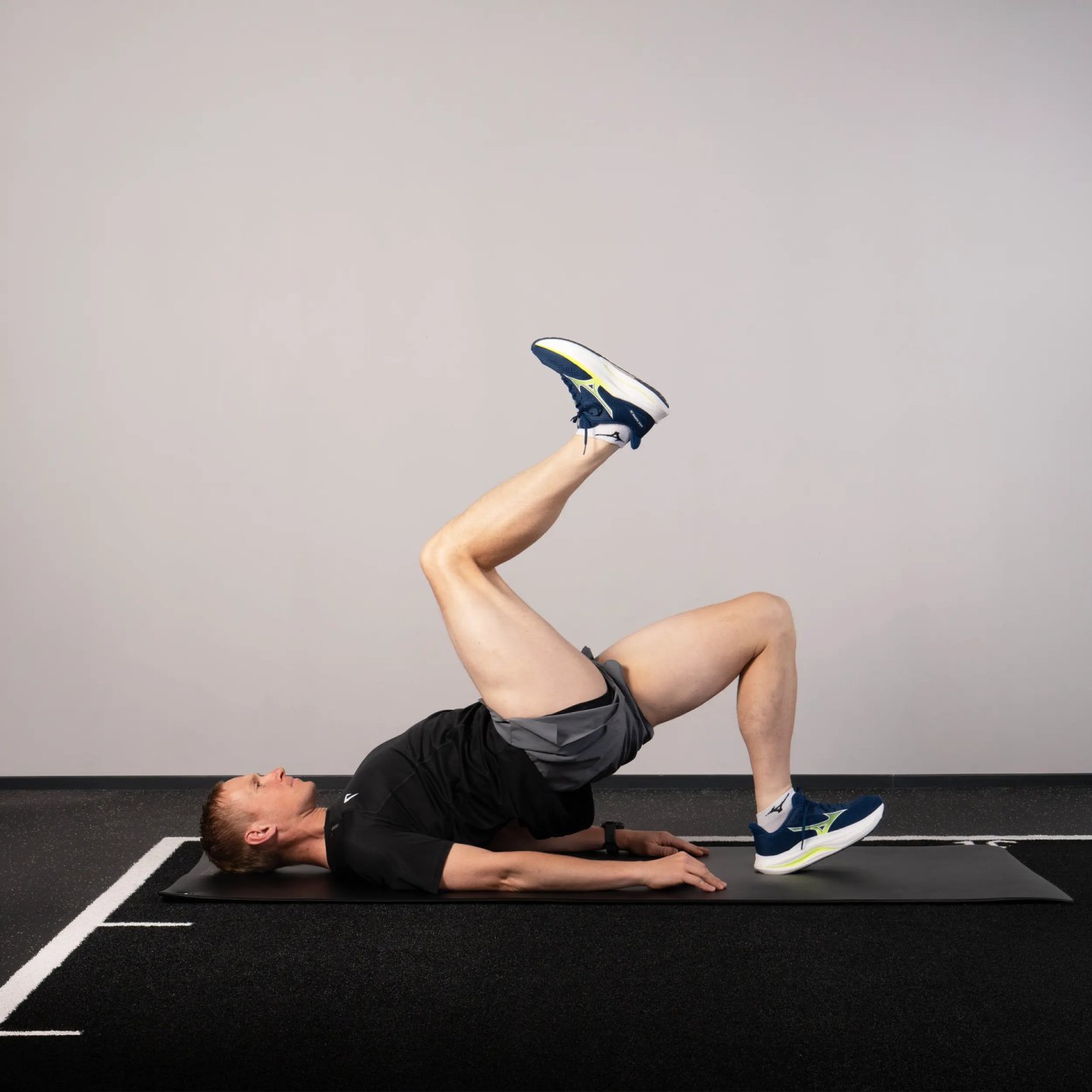 Runner lying on black exercise mat performing a single-leg glute bridge, wearing navy and yellow running shoes and black shorts.
