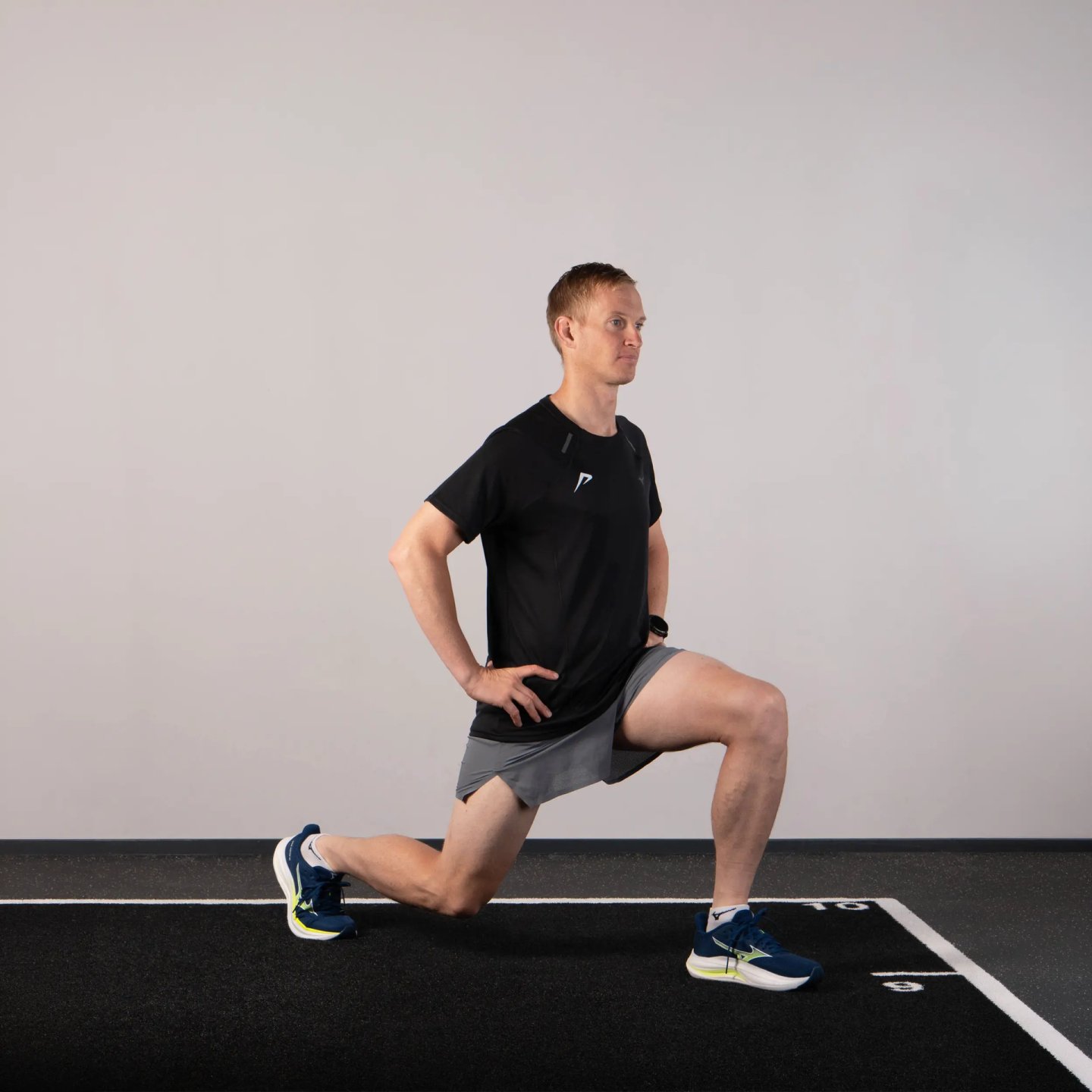 Male runner performing a forward lunge on a gym floor, wearing a black t-shirt, grey shorts and blue running shoes.