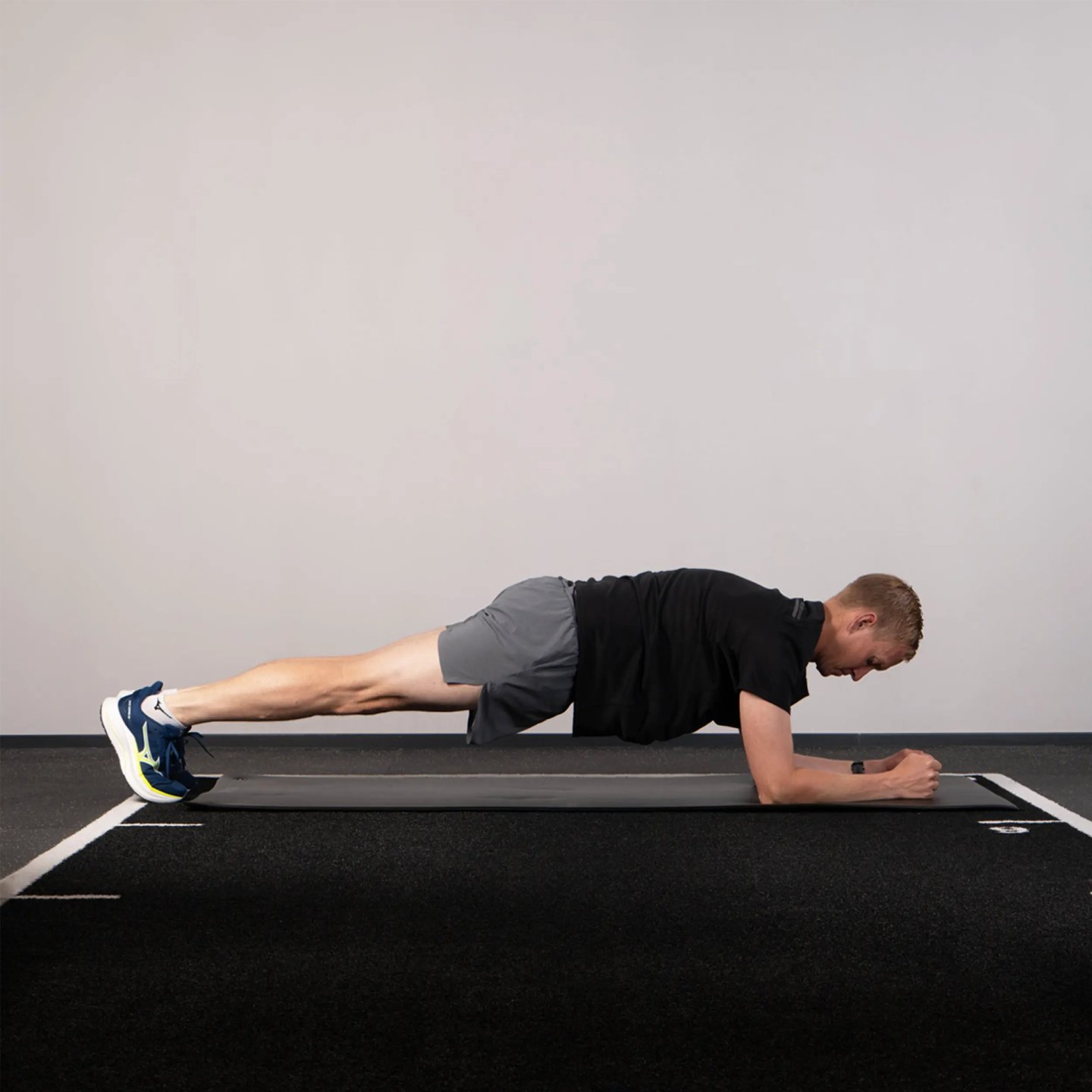 Male runner in black t-shirt and grey shorts performing a forearm plank on a gym mat wearing blue Mizuno running shoes