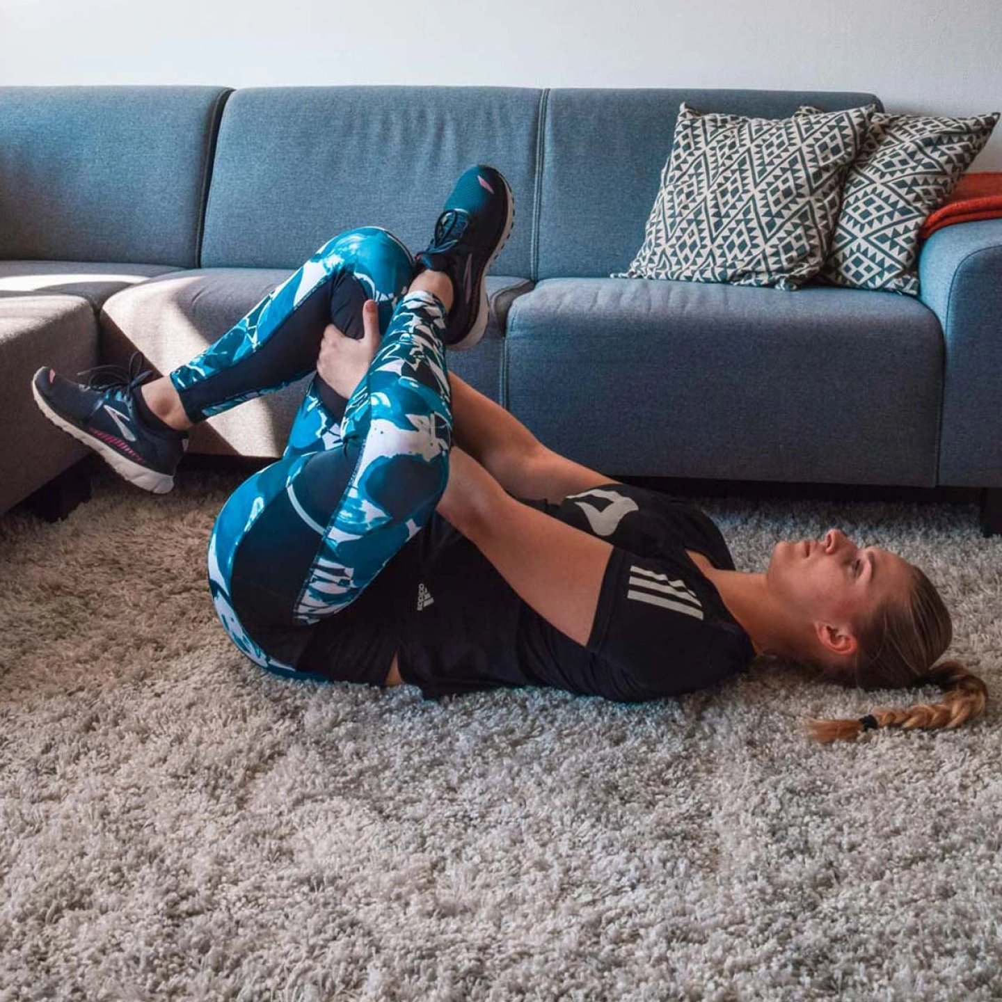 Woman lying on floor in blue patterned leggings and Adidas tee performing a supine knee-to-chest stretch indoors.