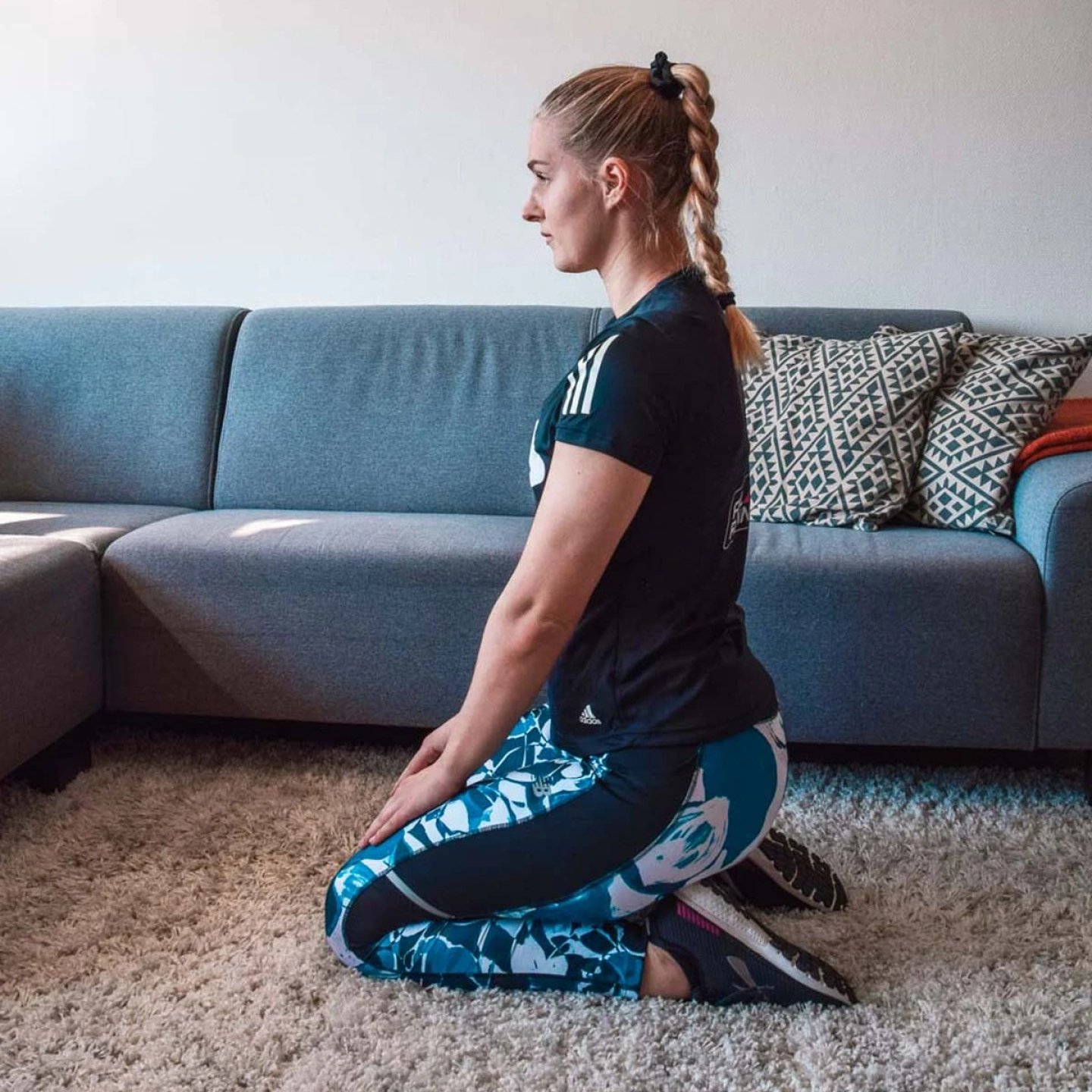 Female runner in Adidas t-shirt and patterned tights kneeling on carpet in a rest stretch position