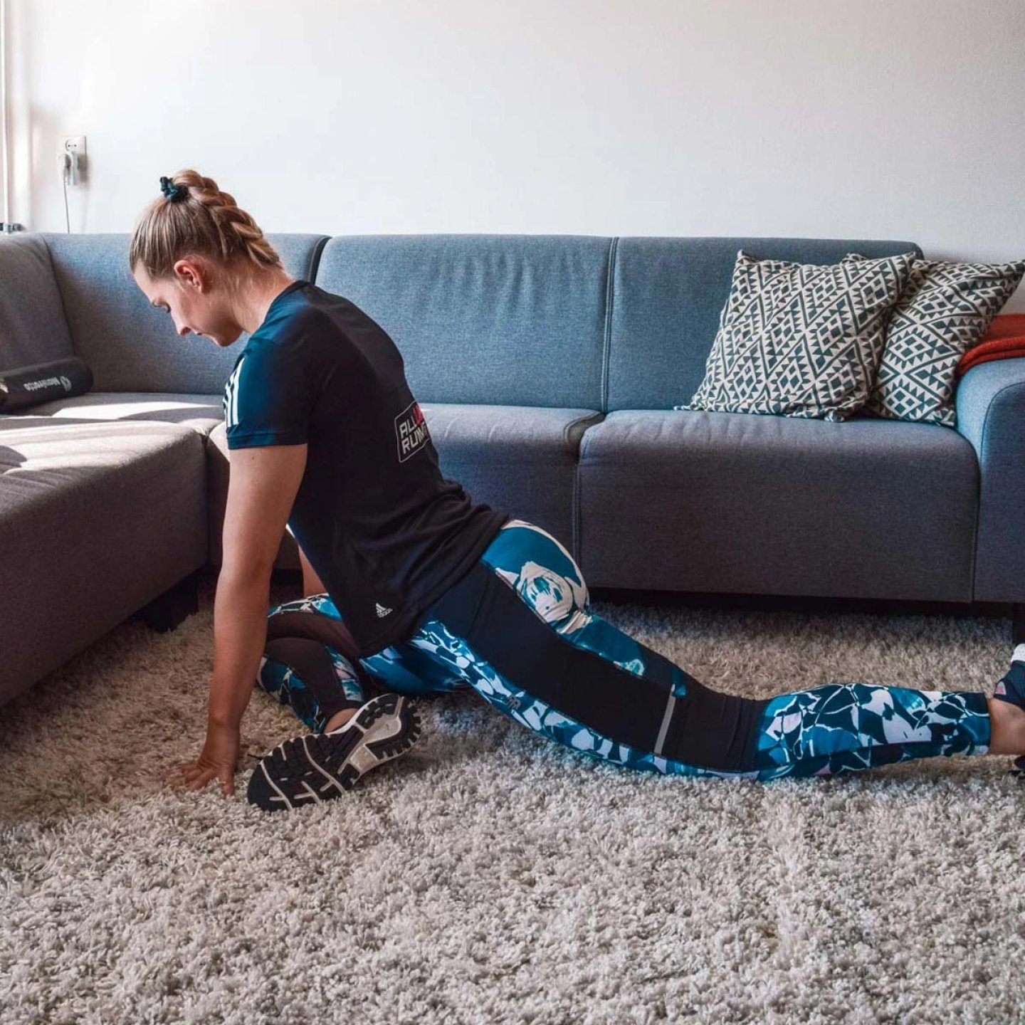 Woman in patterned blue running tights and black training top doing a hip flexor lunge stretch on a carpet at home.
