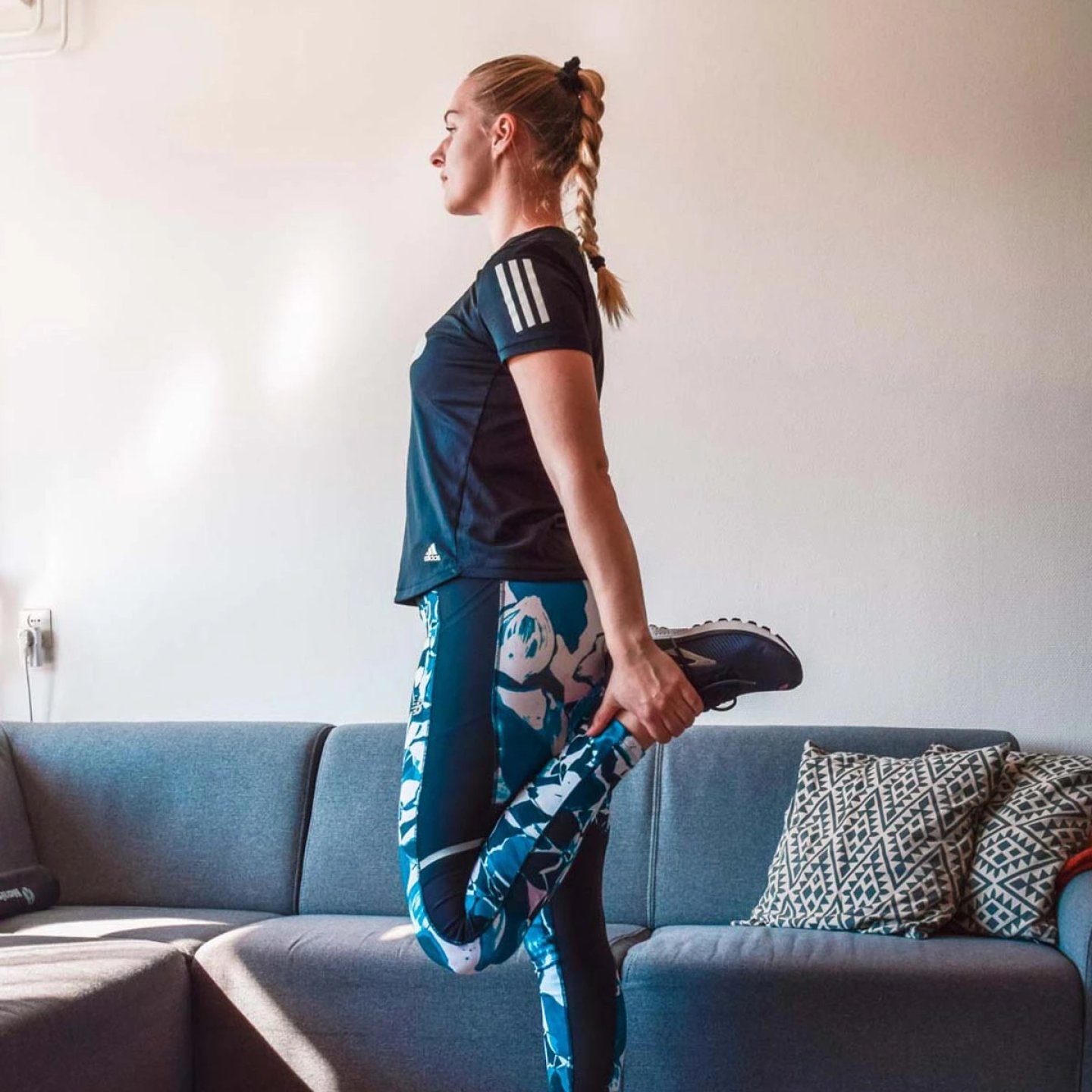 Woman in Adidas running t-shirt and blue printed tights doing a standing quad stretch in a living room