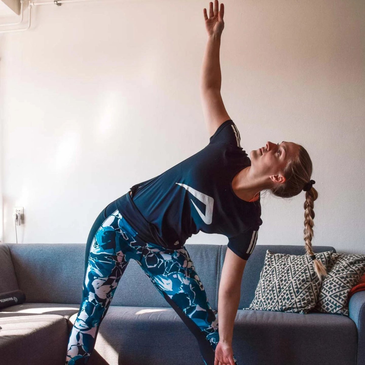 Woman in blue floral running tights and dark top doing a standing triangle stretch in a living room
