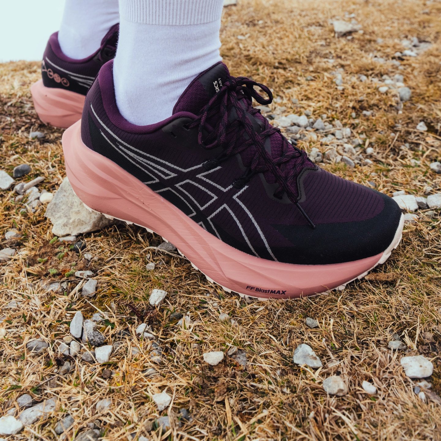 Close-up of Asics FFBlastMAX running shoes in purple and pink on rocky grass terrain, worn with white crew socks.