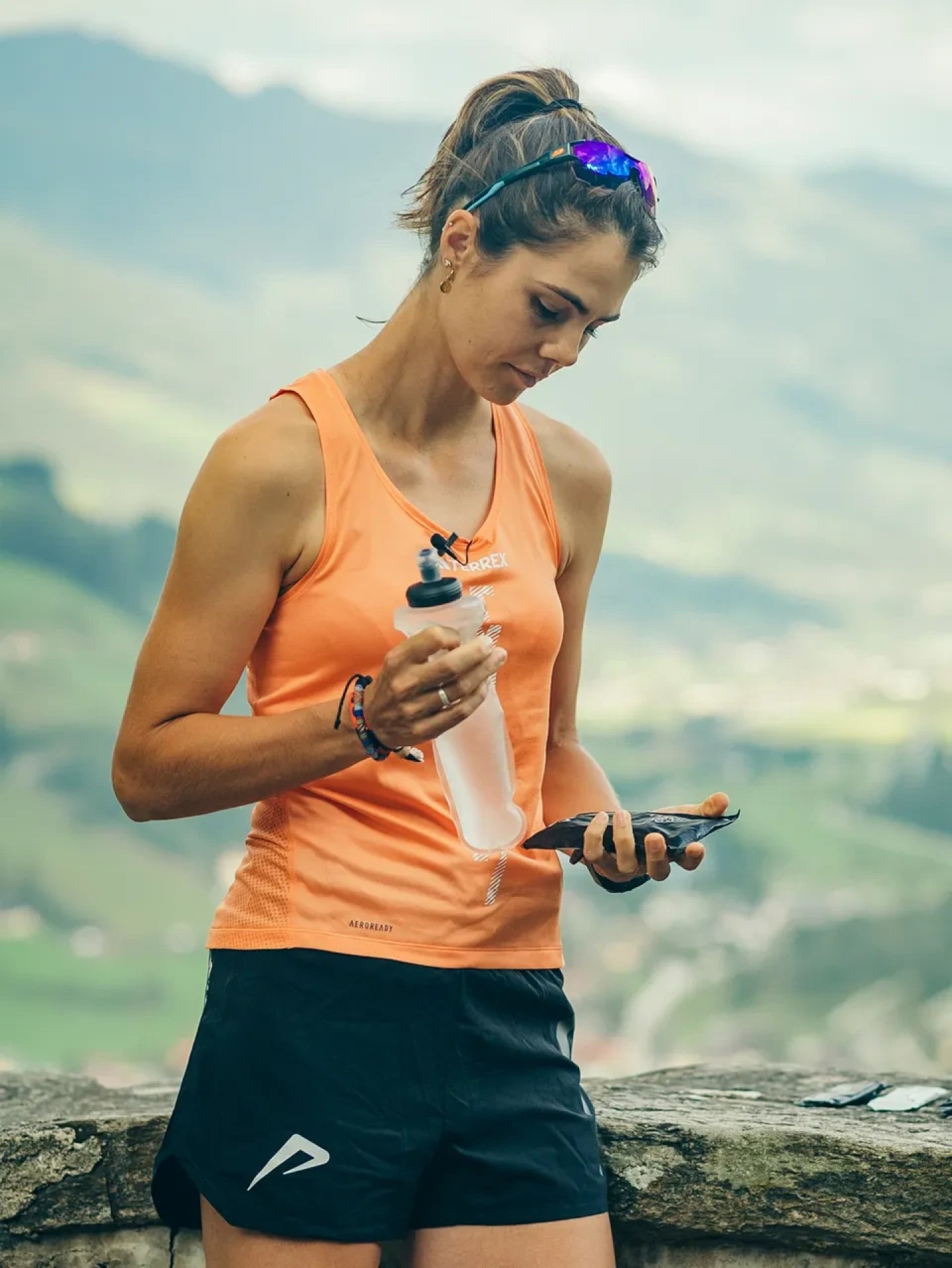 Female trail runner in orange tank top and black shorts holding a soft flask and energy gel at a mountain overlook.