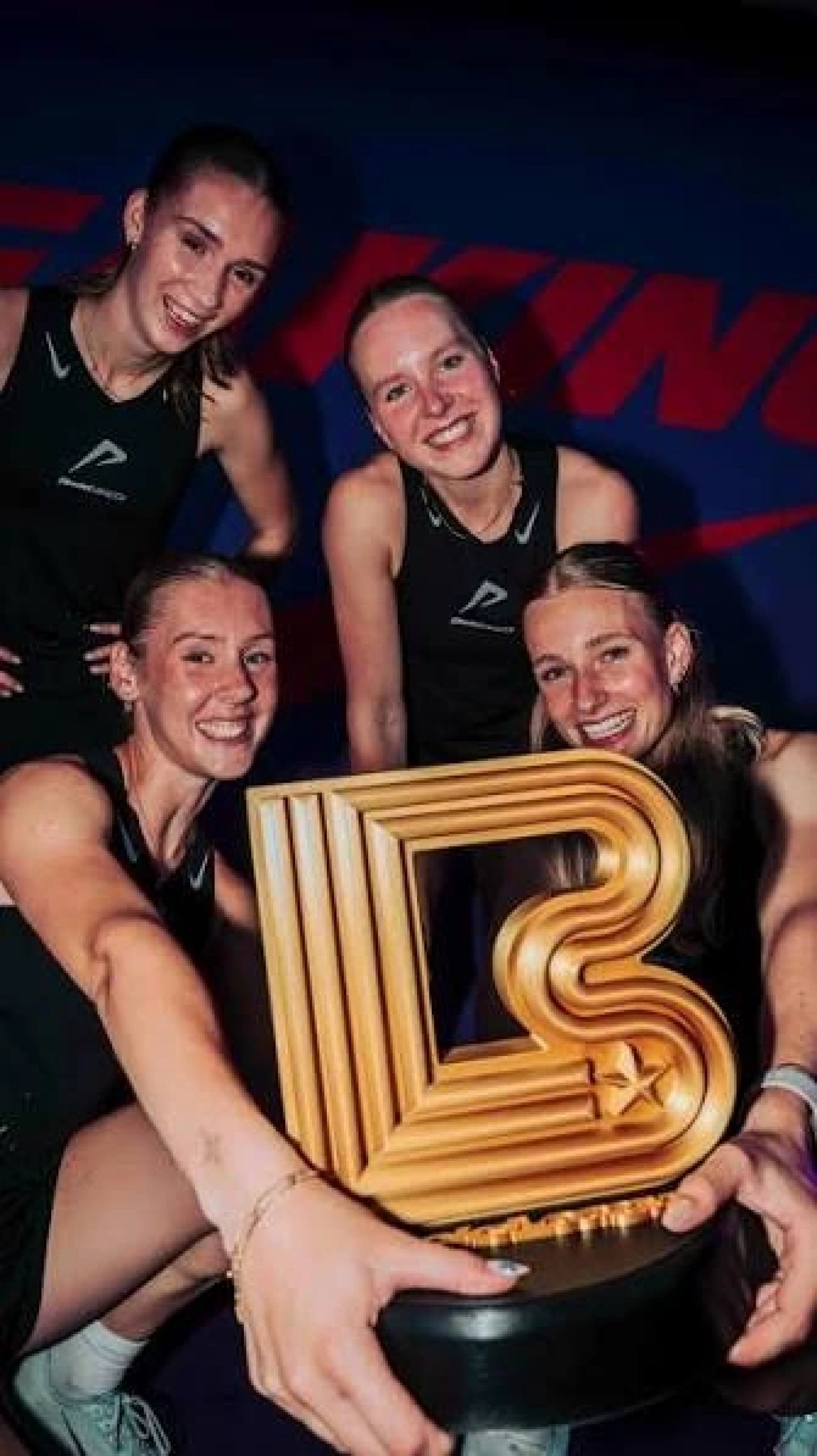 Four smiling female athletes in black athletic vests posing together holding a large gold trophy at an indoor event.