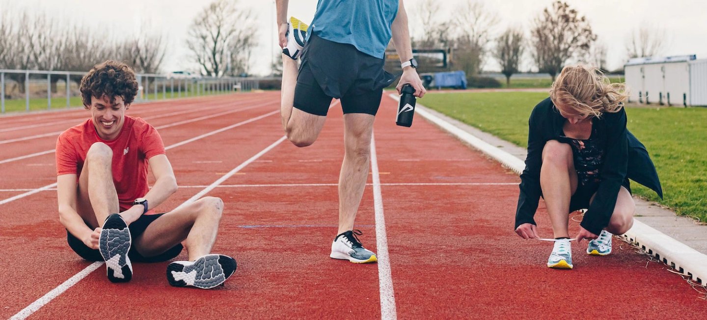 Three runners on a red athletics track tying shoelaces and stretching before a training session, wearing running shoes and sportswear.