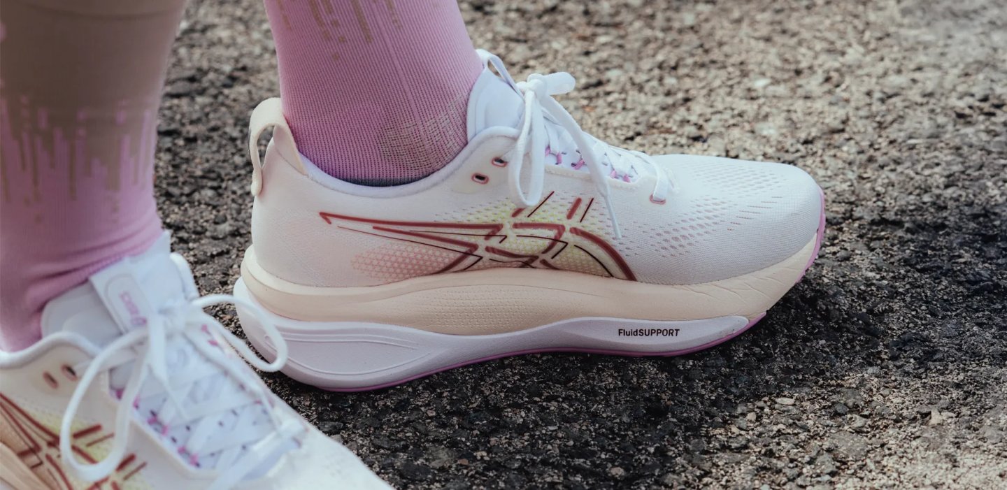 Close-up of a white and pink ASICS running shoe with FluidSupport midsole technology, worn with pink socks on asphalt.