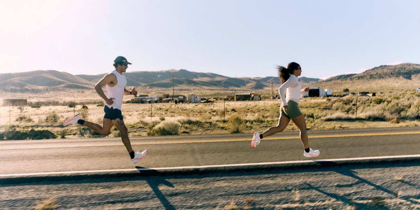 Man and woman running on a desert road wearing white Altra Experience Flow 3 shoes with coral midsoles, arid landscape behind.