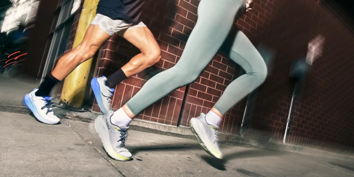 Close-up of two runners wearing Altra FWD Via 2 road running shoes on an urban sidewalk beside a brick wall.