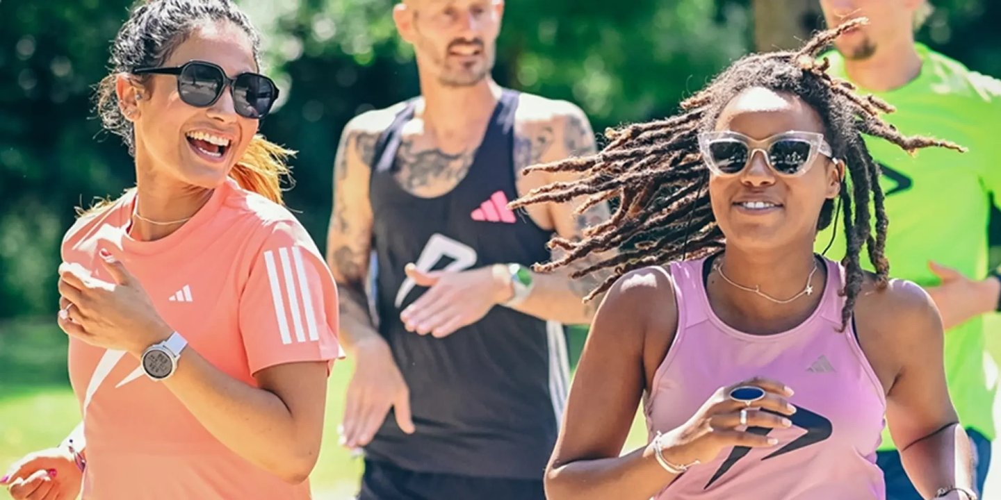 Group of four runners wearing Adidas running apparel jogging together outdoors on a sunny day in a park.