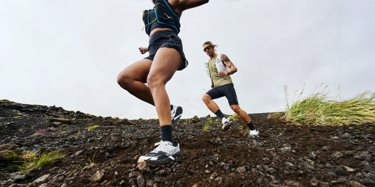 Two trail runners ascending a rocky volcanic slope wearing Hoka Speedgoat 7 trail running shoes and hydration vests.