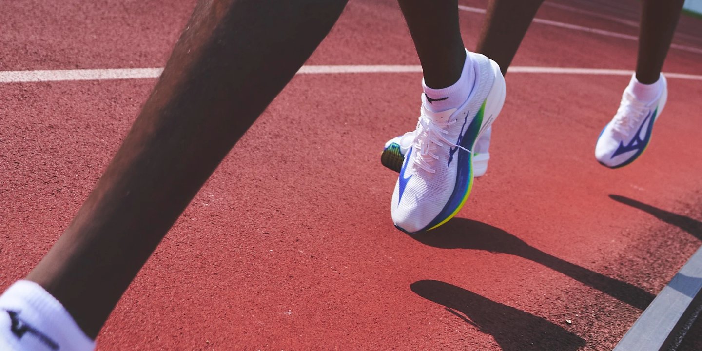 Close-up of two runners wearing white Mizuno Hyperwarp Collective shoes mid-stride on a red running track in sunlight.