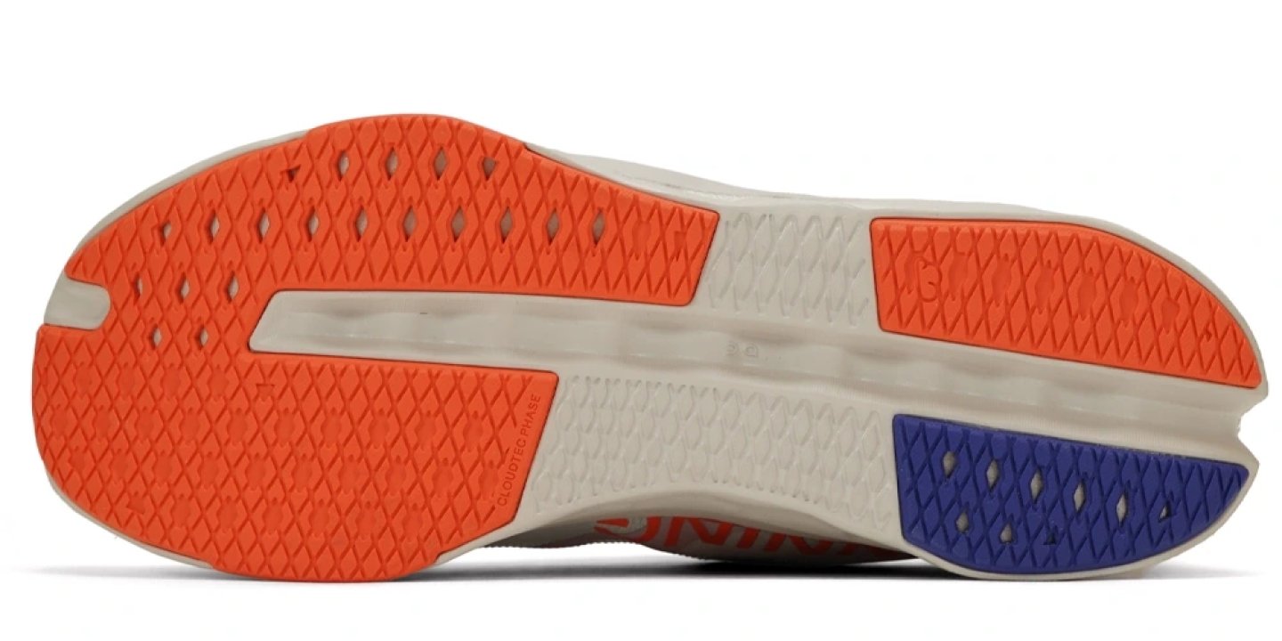 Bottom view of On Cloudsurfer Next running shoe outsole with orange and blue rubber traction zones on a cream midsole