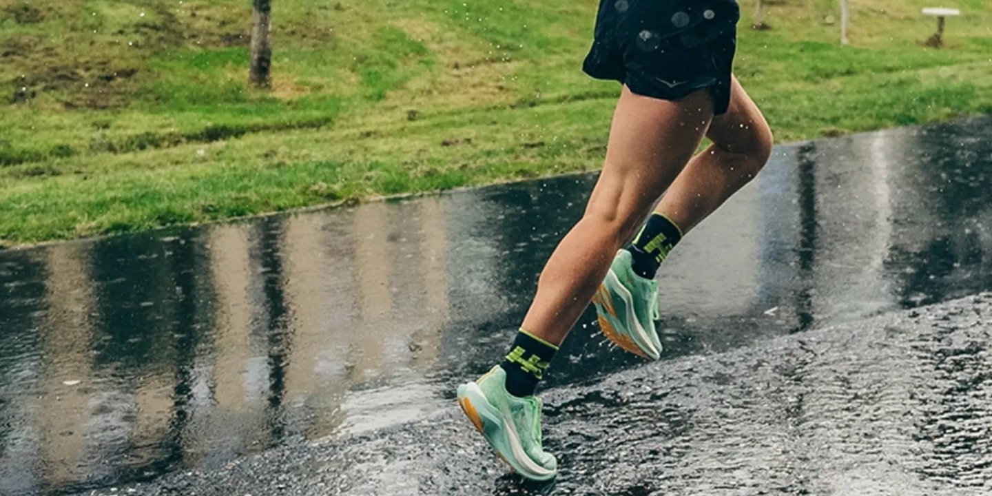 Close-up of a runner's legs mid-stride on a wet road in the rain, wearing mint-green running shoes and black socks.