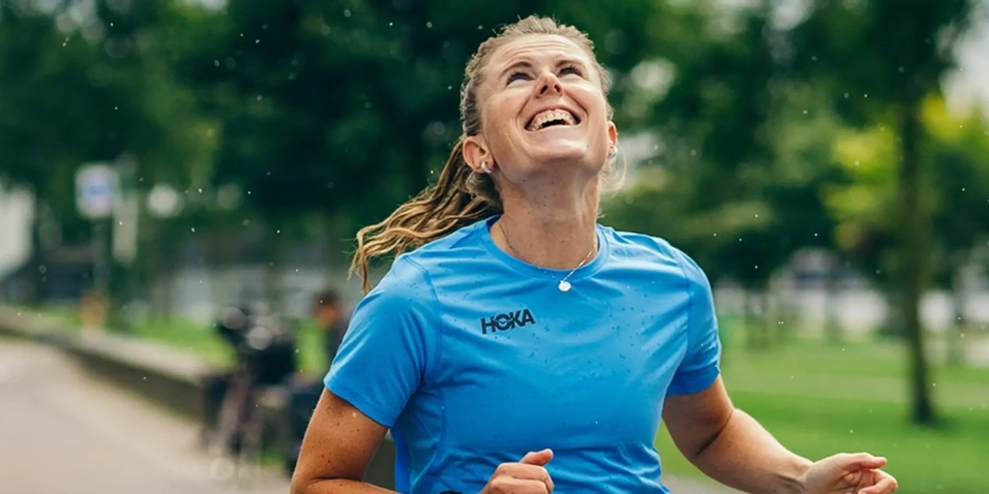 Smiling woman running outdoors in light rain wearing a blue HOKA branded short-sleeve running T-shirt