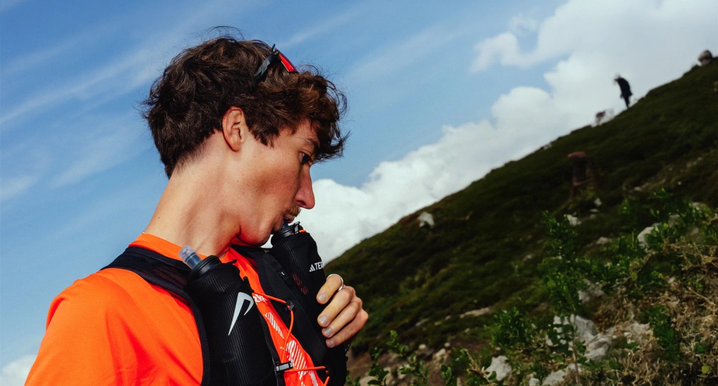 Male trail runner in orange top and black running vest drinking from a soft flask on a green mountain hillside