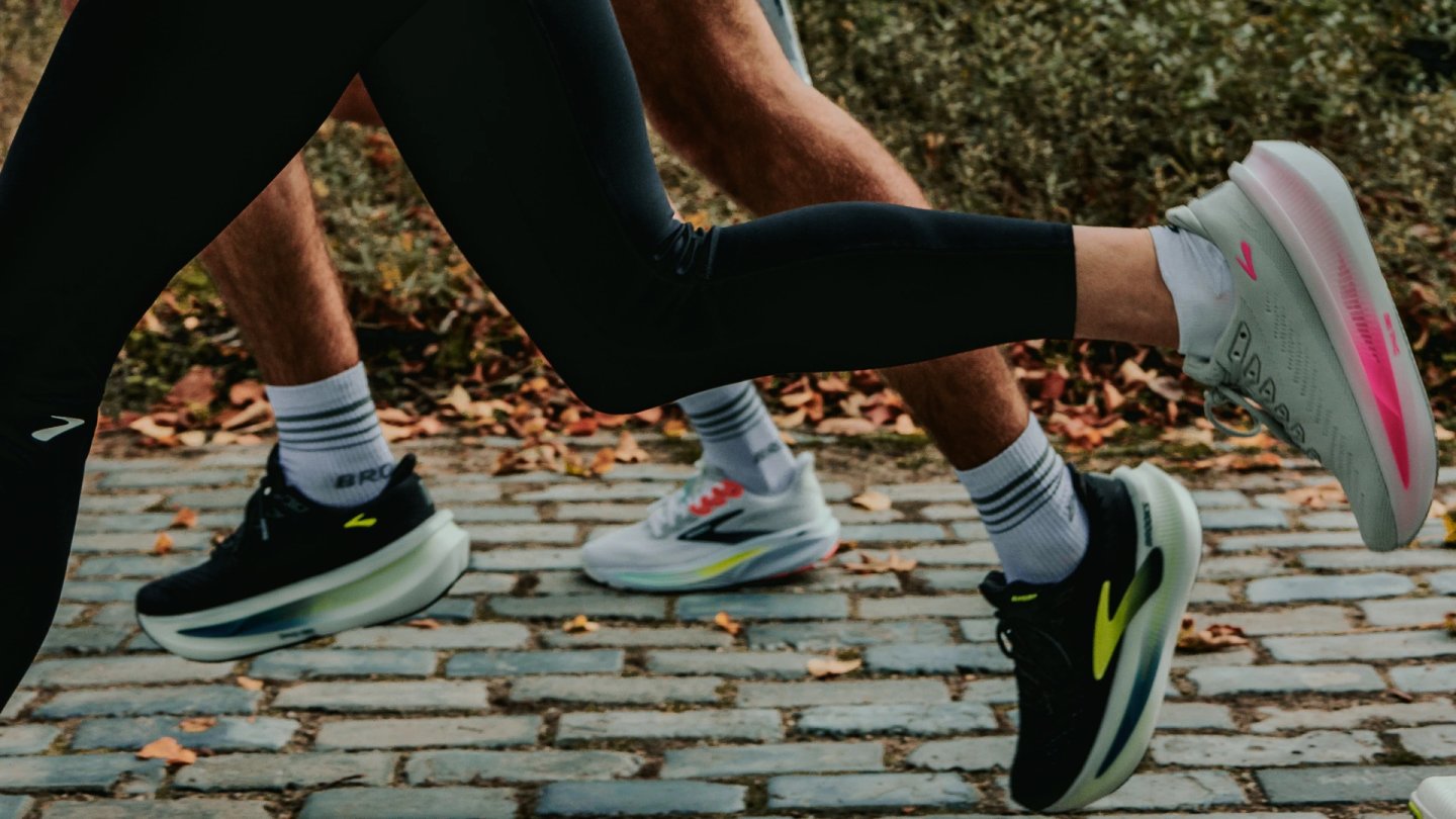 Close-up of three pairs of Brooks running shoes worn by runners on a cobblestone path covered with autumn leaves.