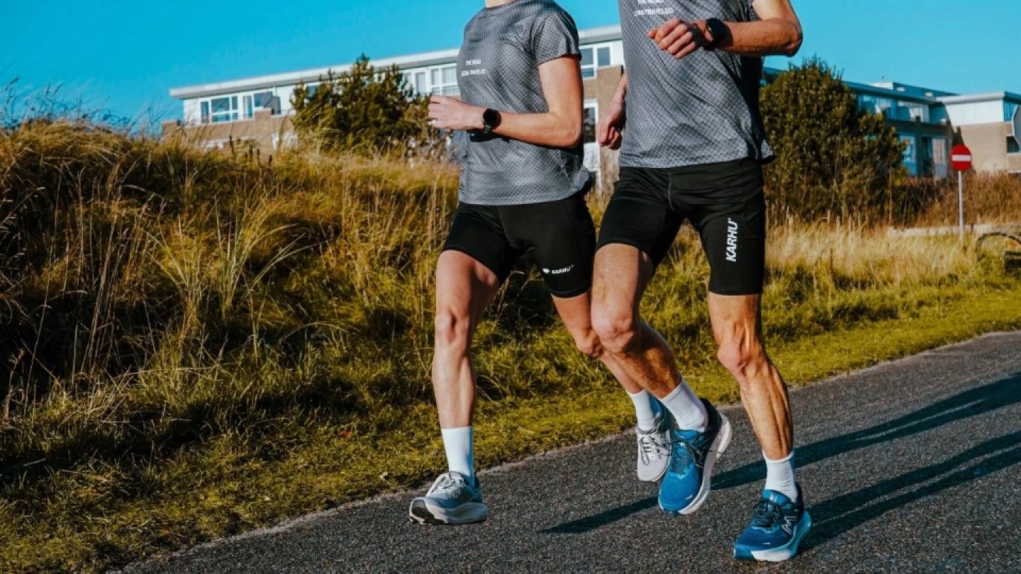 Two male runners in black Karhu shorts and grey t-shirts running on a road with golden grass and blue sky behind them.