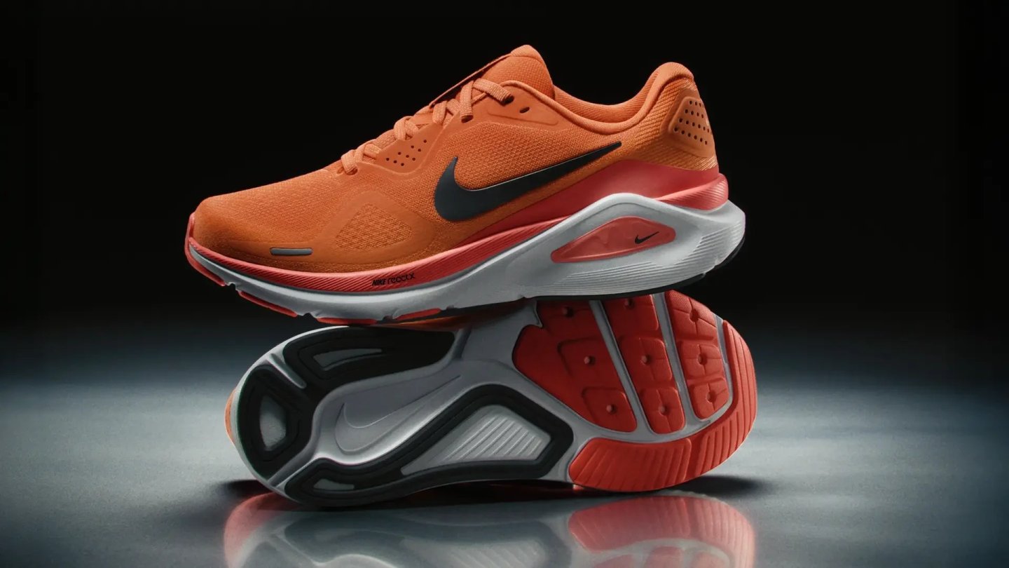 Nike Structure 26 running shoe in orange and red, displayed sole-up and side-on against a dark studio background.