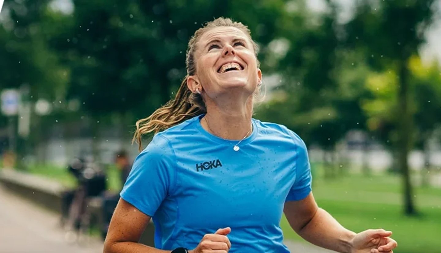 Smiling woman running in a blue HOKA branded short-sleeve running t-shirt through a park in light rain
