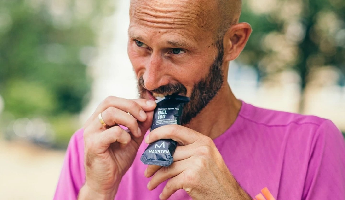 Bearded male runner in a pink t-shirt tearing open a Maurten Gel 100 energy gel packet with his teeth outdoors.