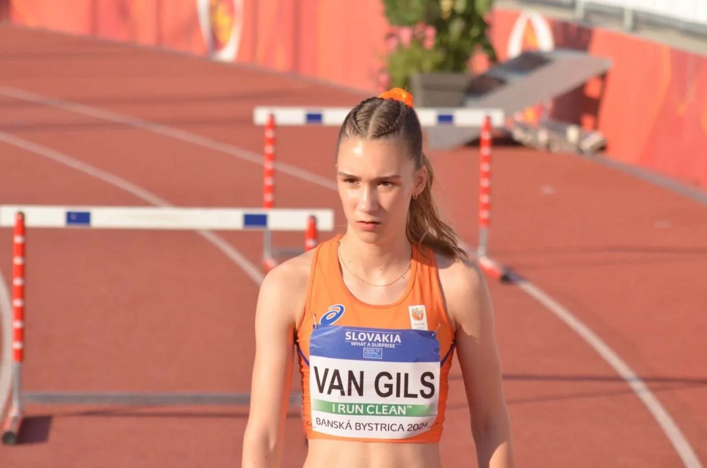Athlete Van Gils in orange Netherlands Asics singlet at Banská Bystrica 2024 athletics meet, hurdles visible in background.