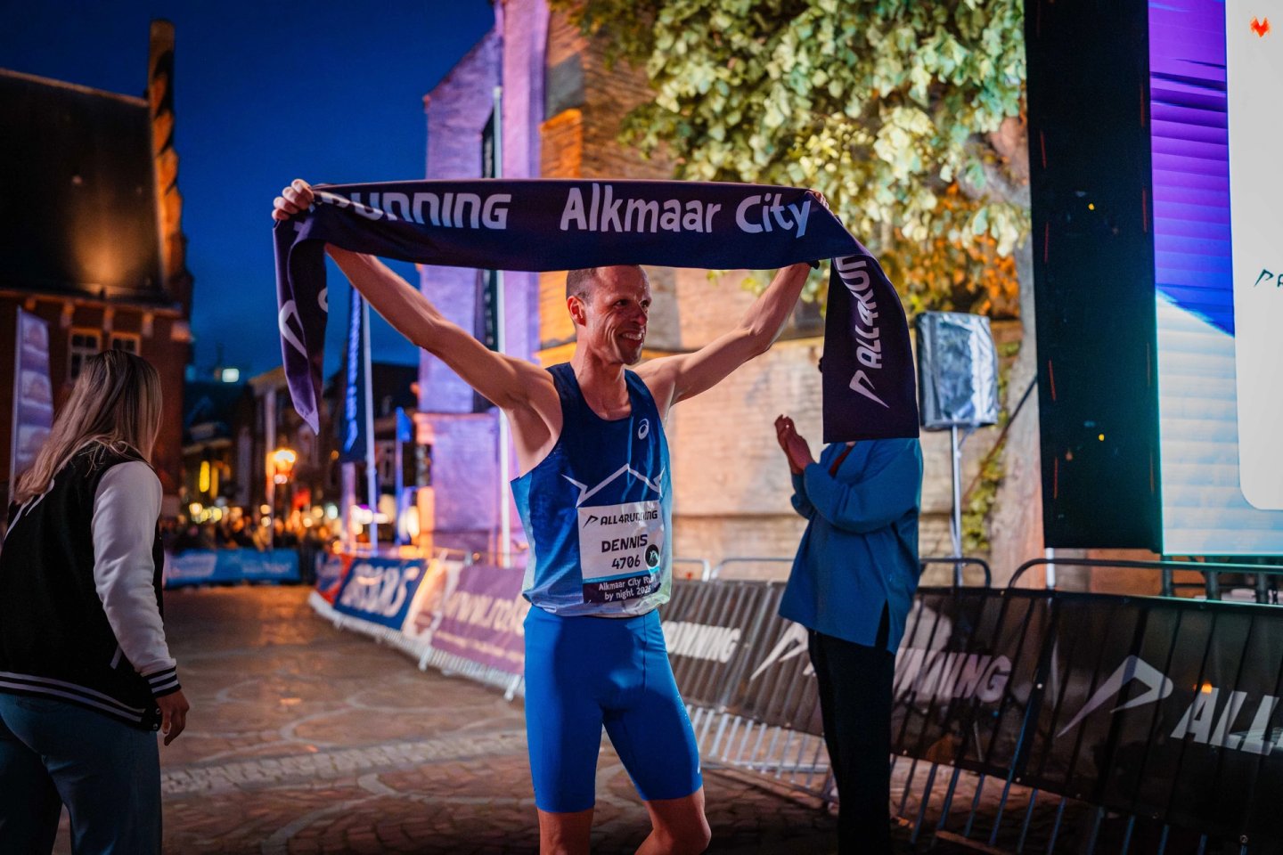 Male runner named Dennis, bib 4706, celebrates at the Alkmaar City Run by Night finish line holding up a race banner.