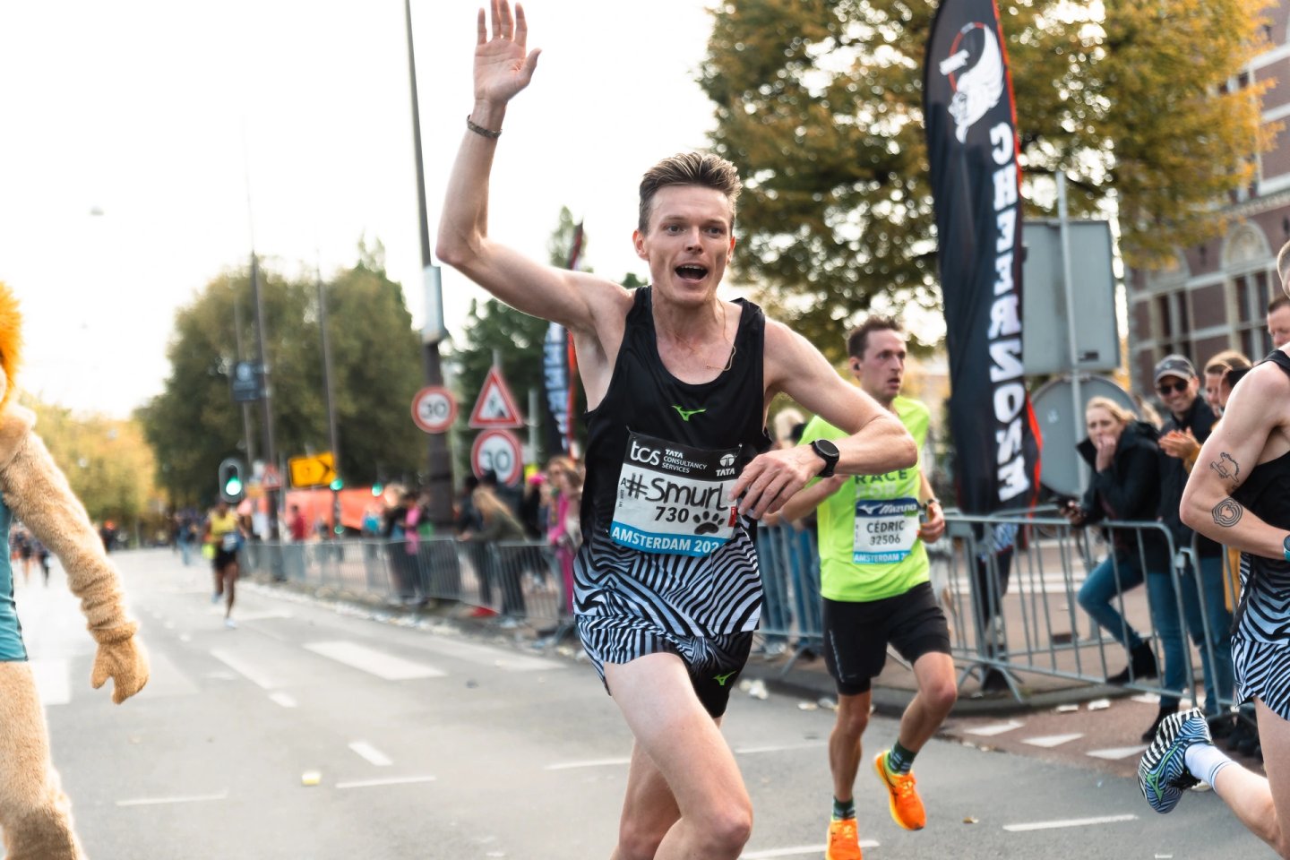 Male marathon runner bib 730 waving hand mid-race at TCS Amsterdam Marathon wearing black Mizuno vest and zebra-print shorts.