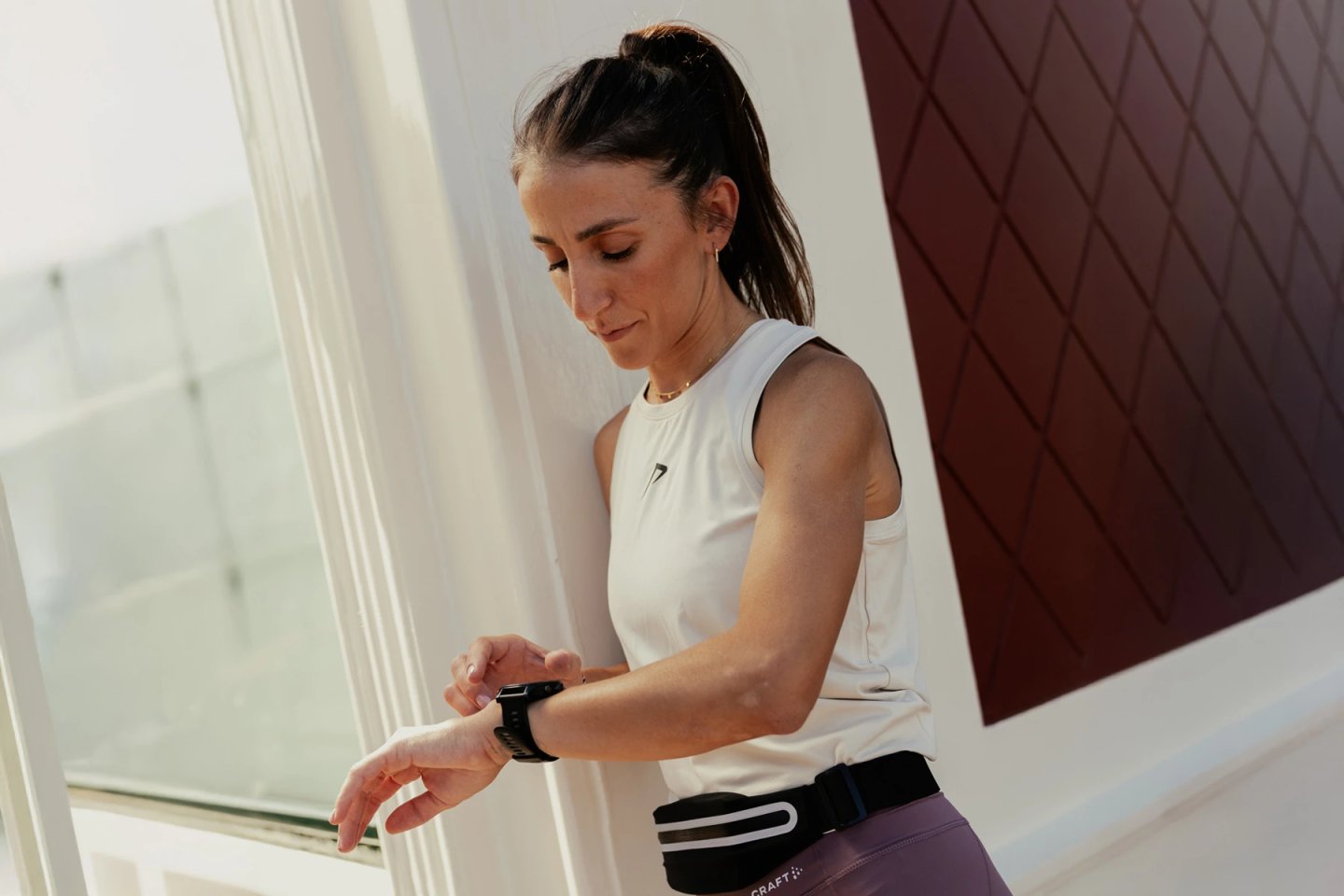 Female runner in white sleeveless top and purple Craft tights checking a black GPS sports watch on her wrist, wearing a running belt