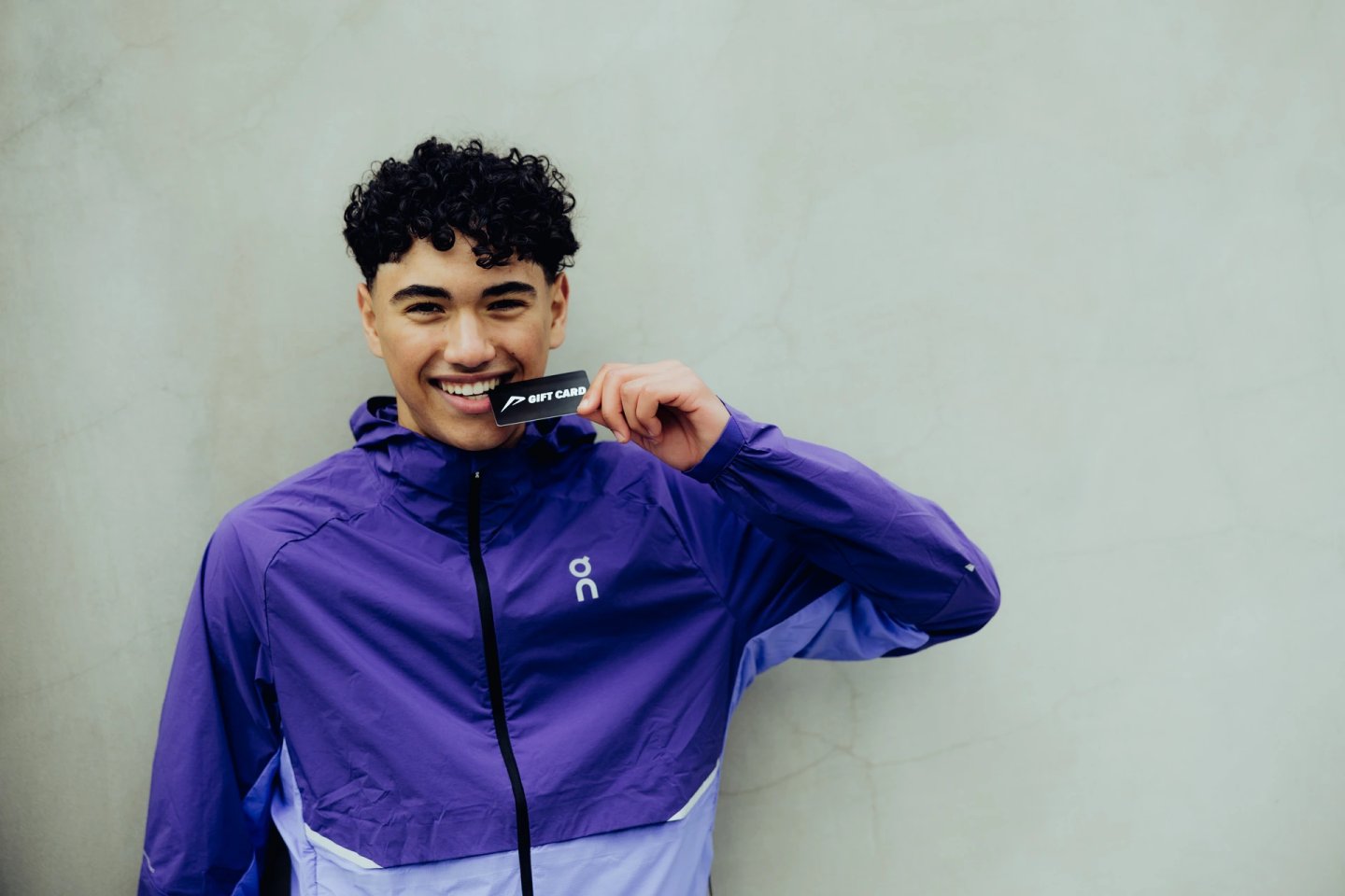 Smiling young man wearing a purple On Running jacket holding a black gift card close to his face against a grey wall.