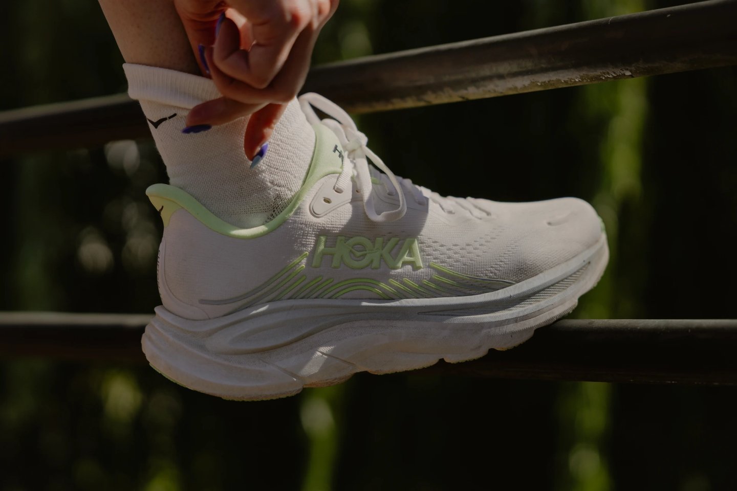 Close-up of a white HOKA running shoe with lime green branding and heel collar, worn with white ankle socks.