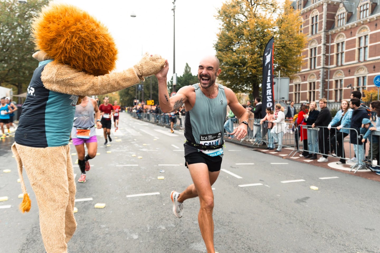 Smiling bald male marathon runner in grey On vest and black shorts high-fiving a lion mascot during a city marathon race.