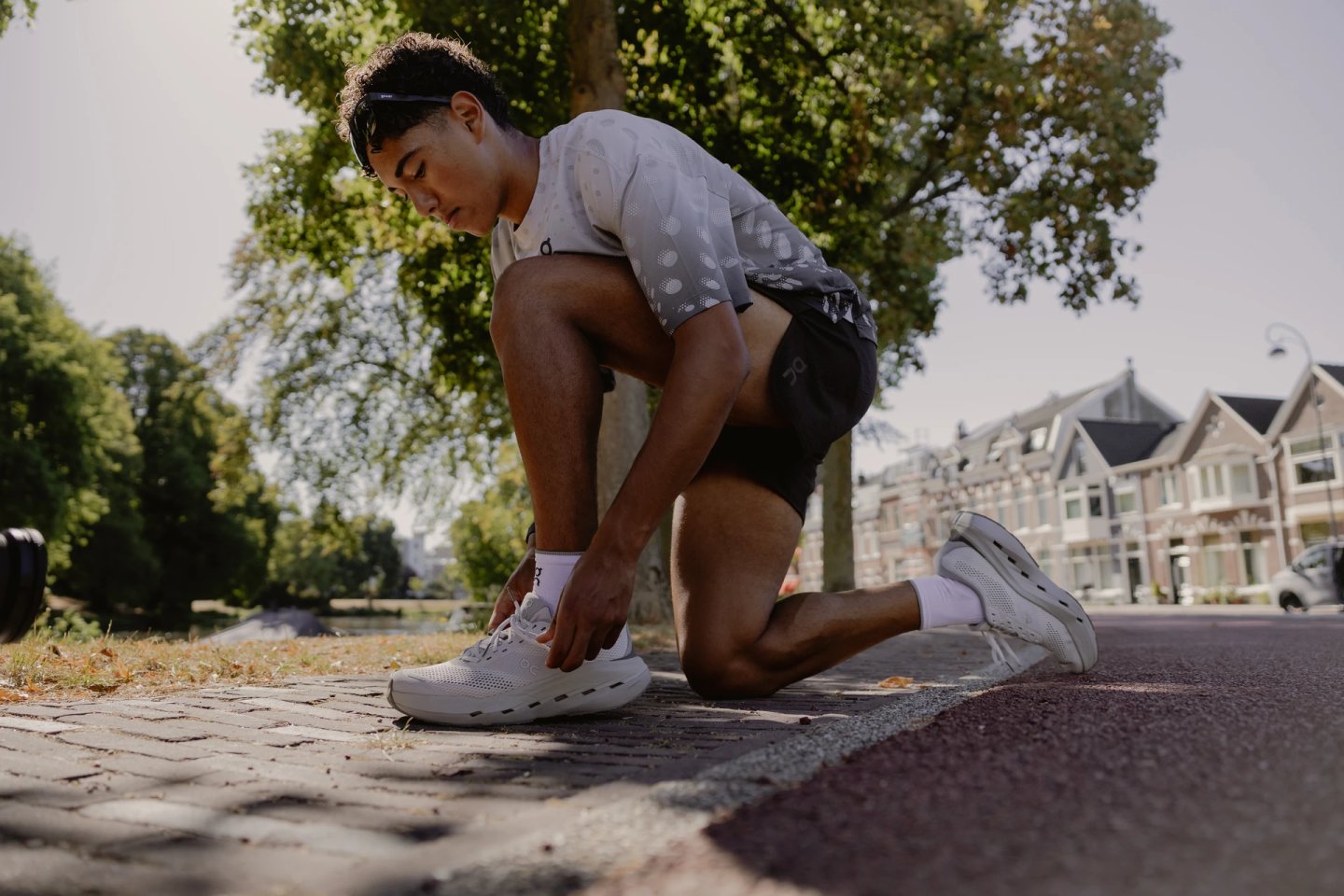 Male runner kneeling on a cobblestone path tying laces on white On running shoes, wearing grey graphic tee and black shorts