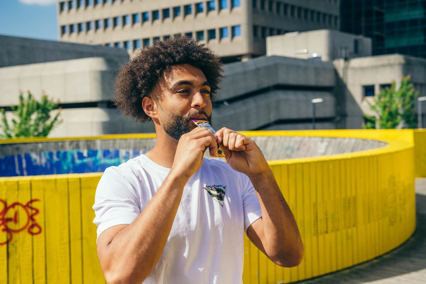 Male runner in white mesh t-shirt consuming a SIS energy gel outdoors in an urban setting with a yellow wall behind him.