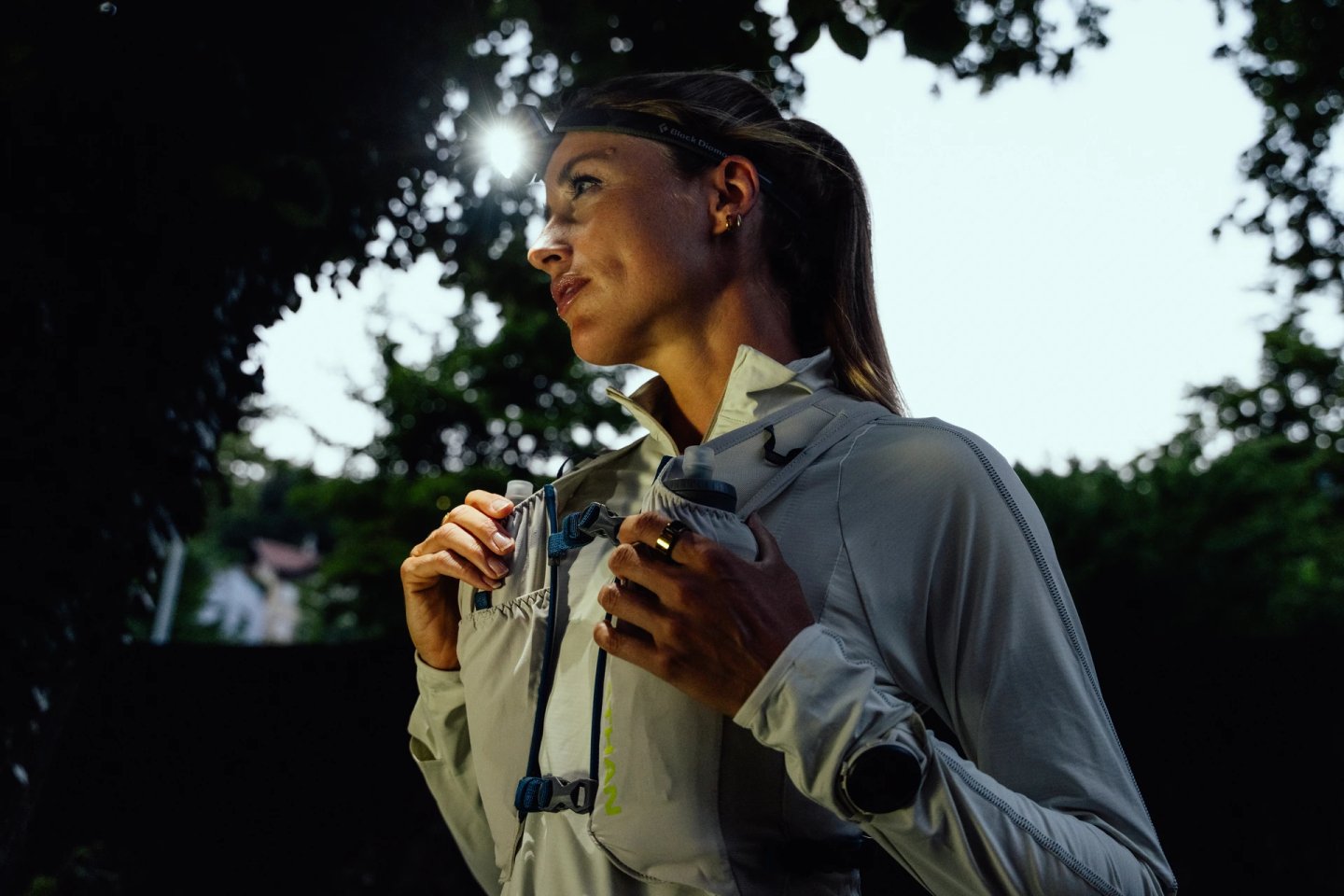 Female trail runner wearing a Black Diamond headlamp and light tan running vest with blue buckles, running outdoors at dusk or dawn.