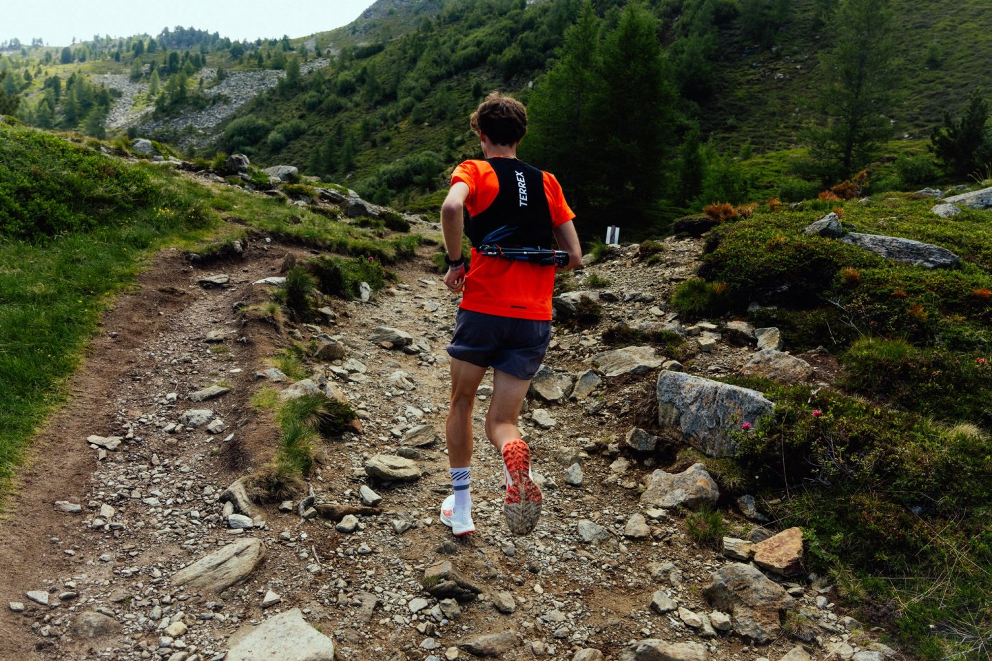 Male trail runner from behind ascending a rocky alpine trail in Innsbruck, wearing orange shirt, black Terrex vest and trail shoes.
