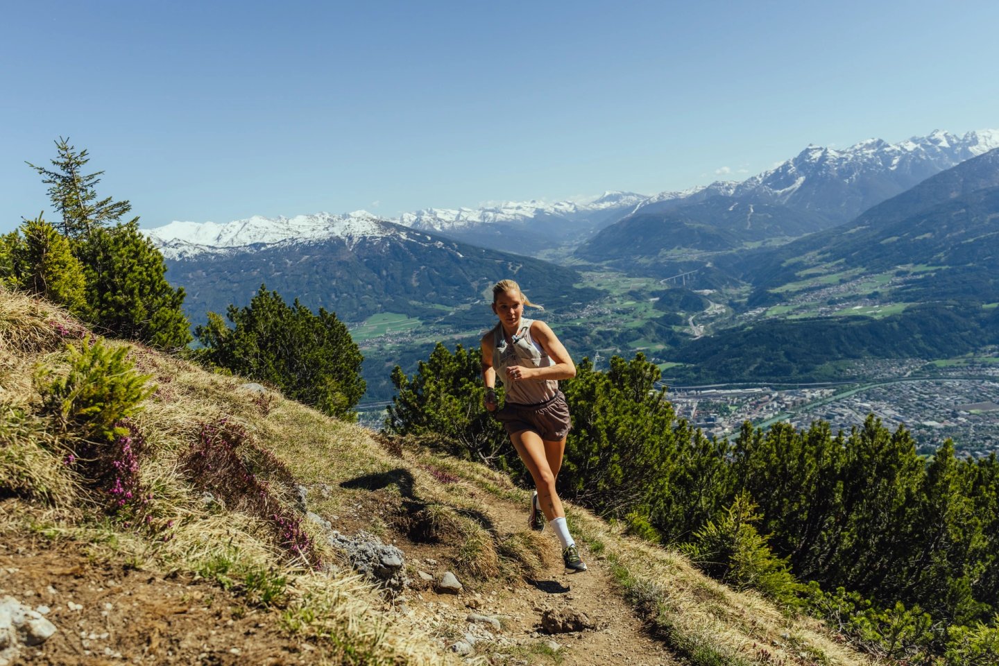 Woman in tank top and shorts trail running uphill on a rocky Alpine path with snow-capped mountains in the background