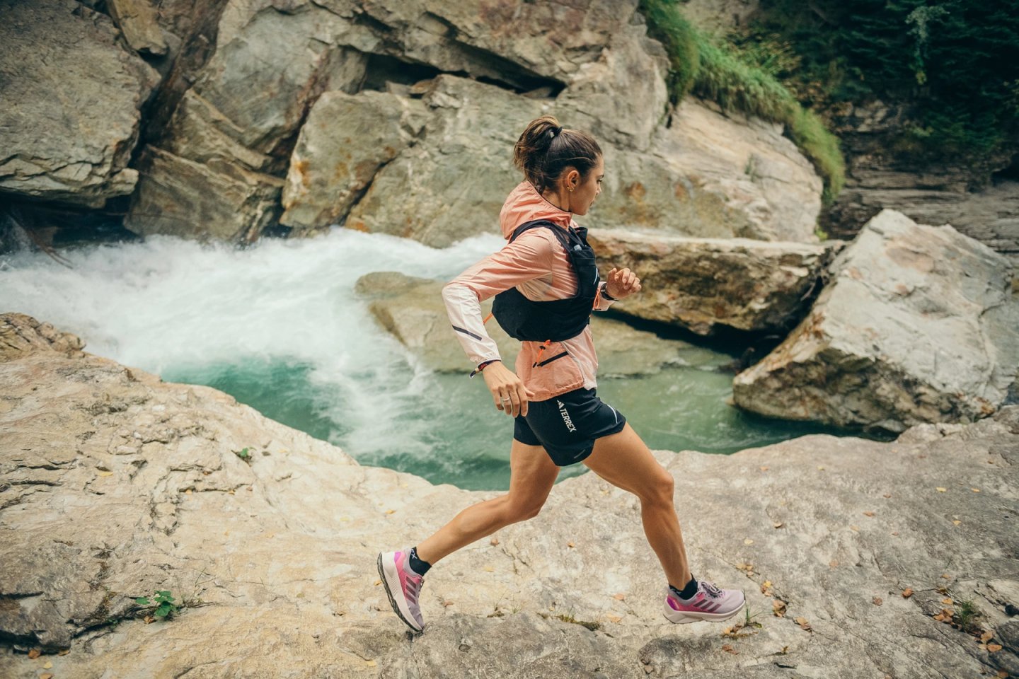 Woman trail running over rocks beside a turquoise waterfall gorge, wearing pink jacket, black shorts, hydration vest and pink running shoes.