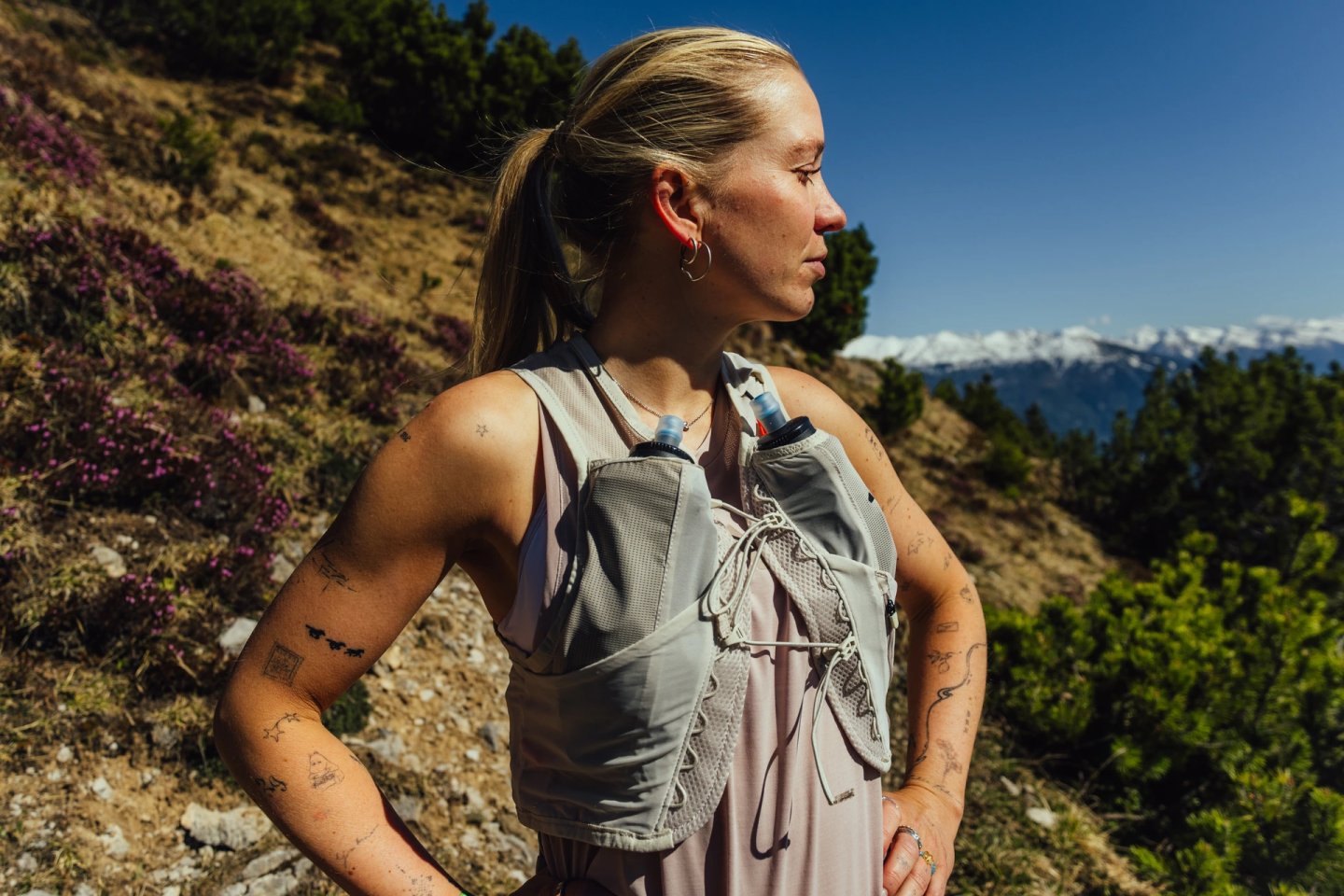 Female trail runner wearing a light grey hydration vest with dual soft flasks on a sunny alpine mountain trail
