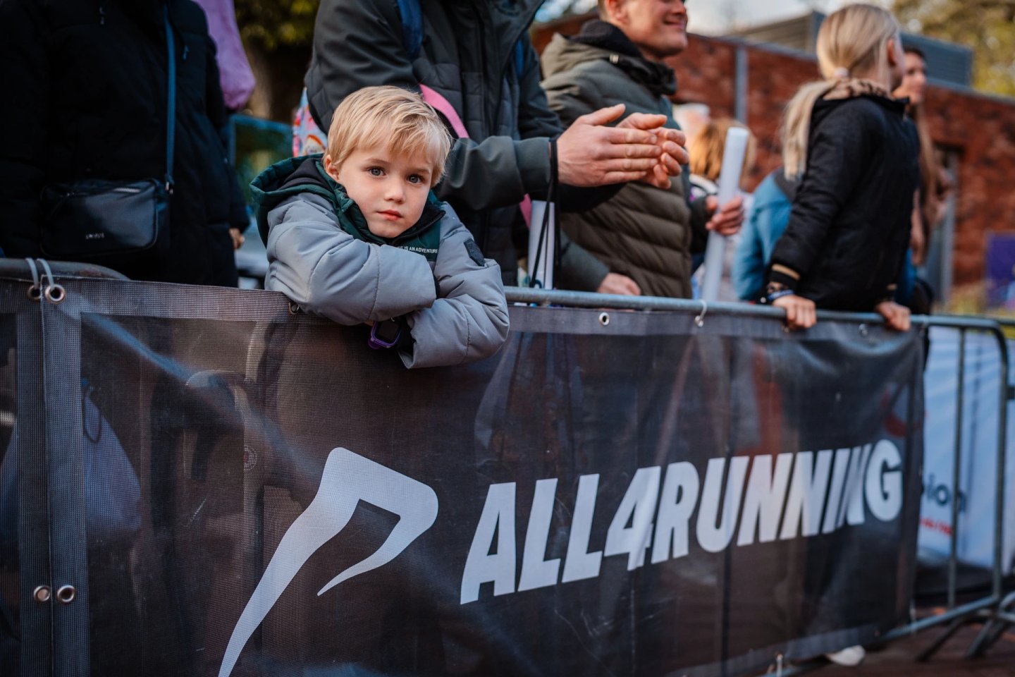 Blonde toddler leaning over a metal crowd barrier at an outdoor running event, spectators clapping in background.