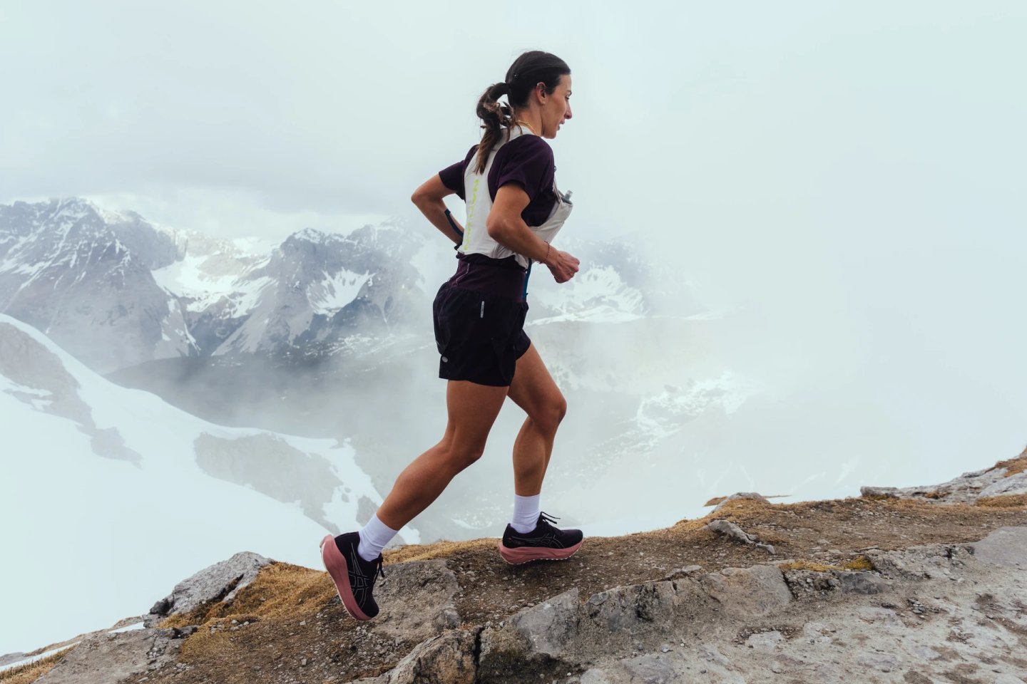Woman trail running on rocky mountain ridge wearing Asics shoes, dark shorts, white hydration vest, snowy peaks in background.