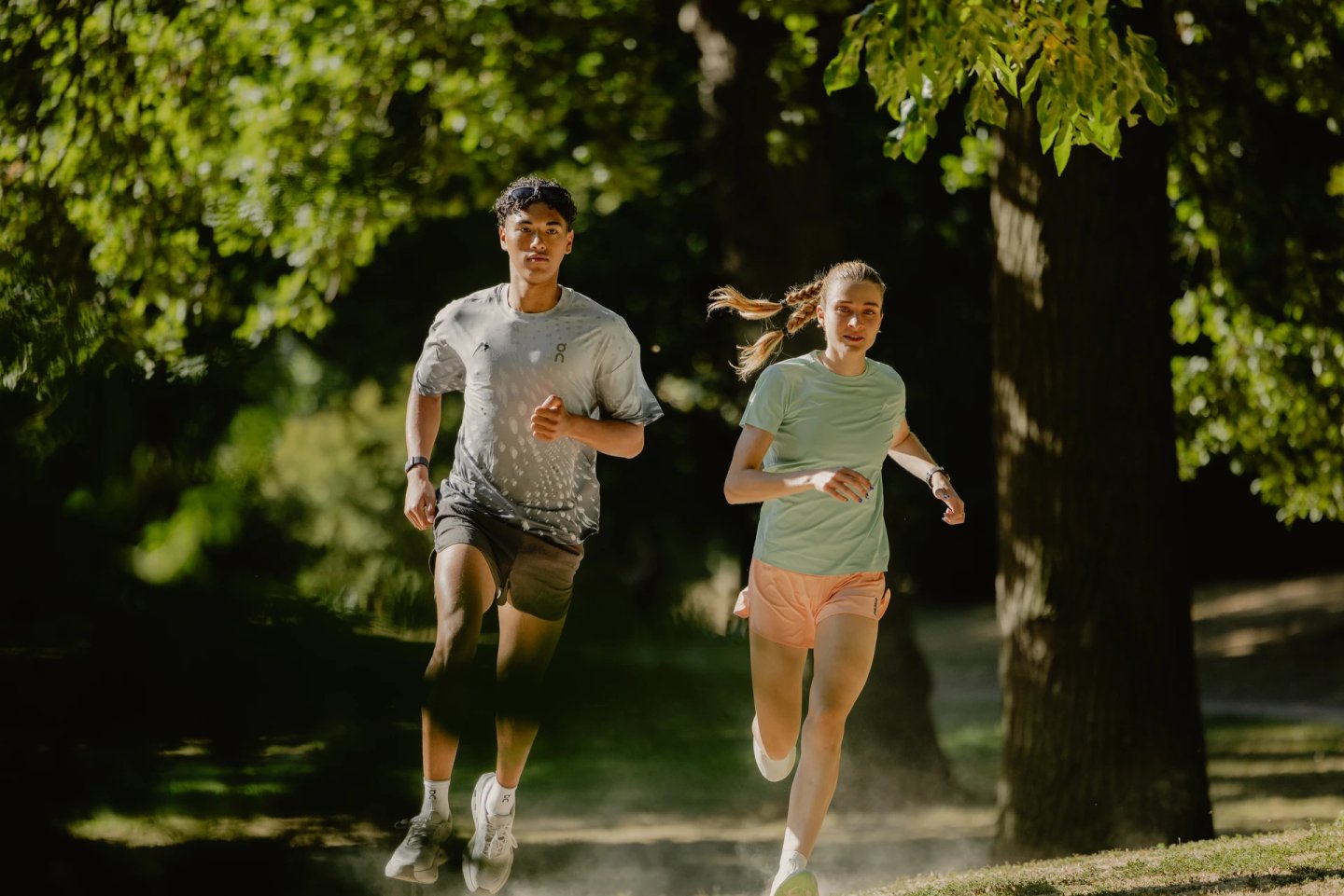 Man in grey graphic tee and dark shorts and woman in mint tee and peach shorts running on a shaded park path in summer.